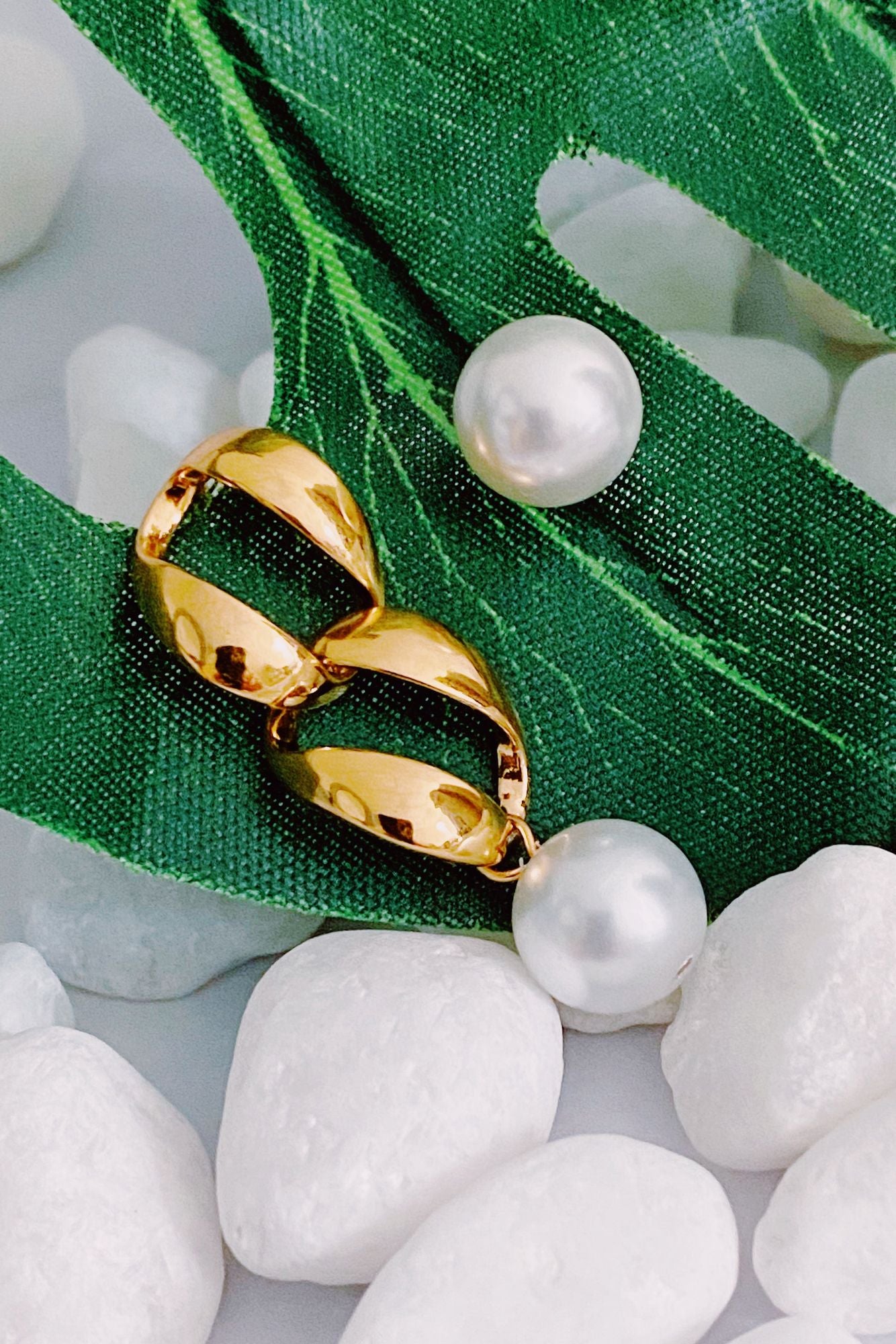 Gold and pearl drop earrings resting on green fabric and white pebbles, showcasing a modern design with a shiny gold finish and elegant round pearls.