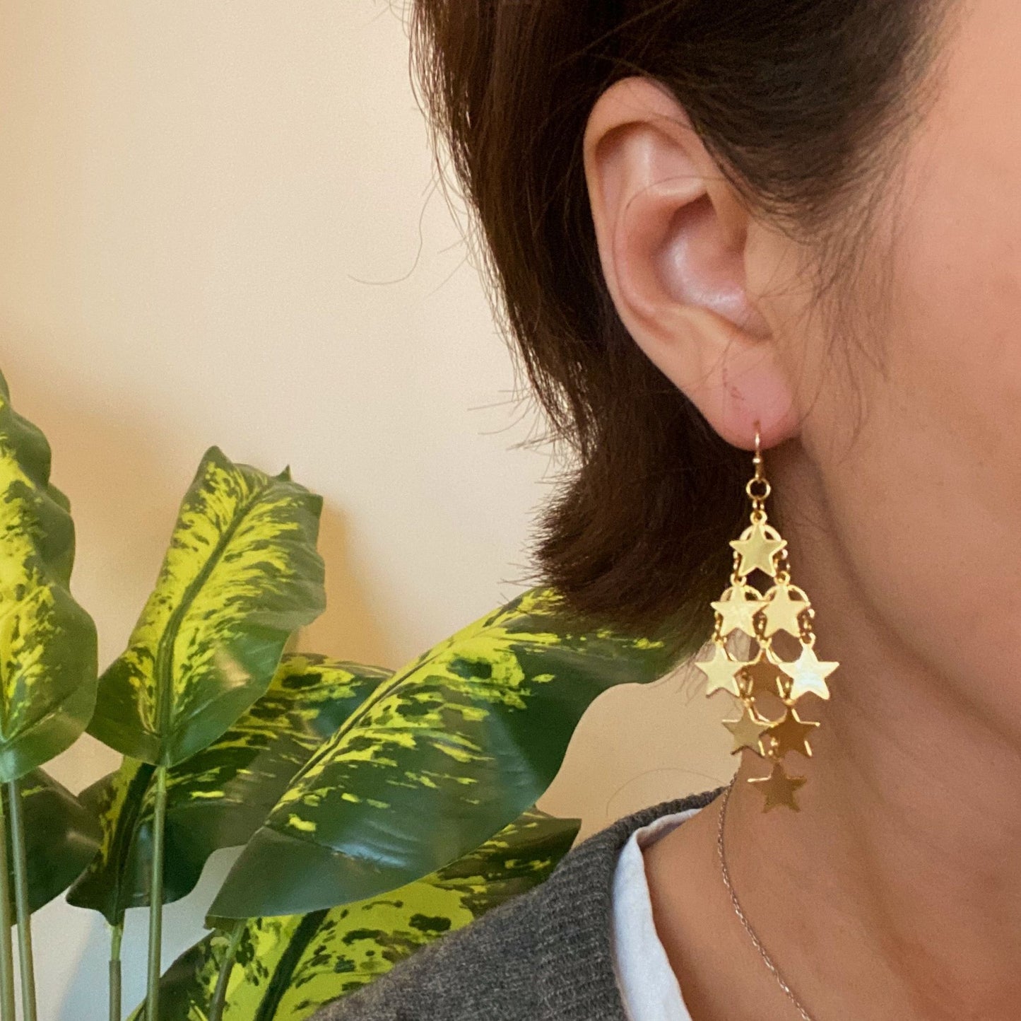 Gold star-shaped dangle earrings worn by a woman, showcasing a stylish design against a background of green plant leaves.