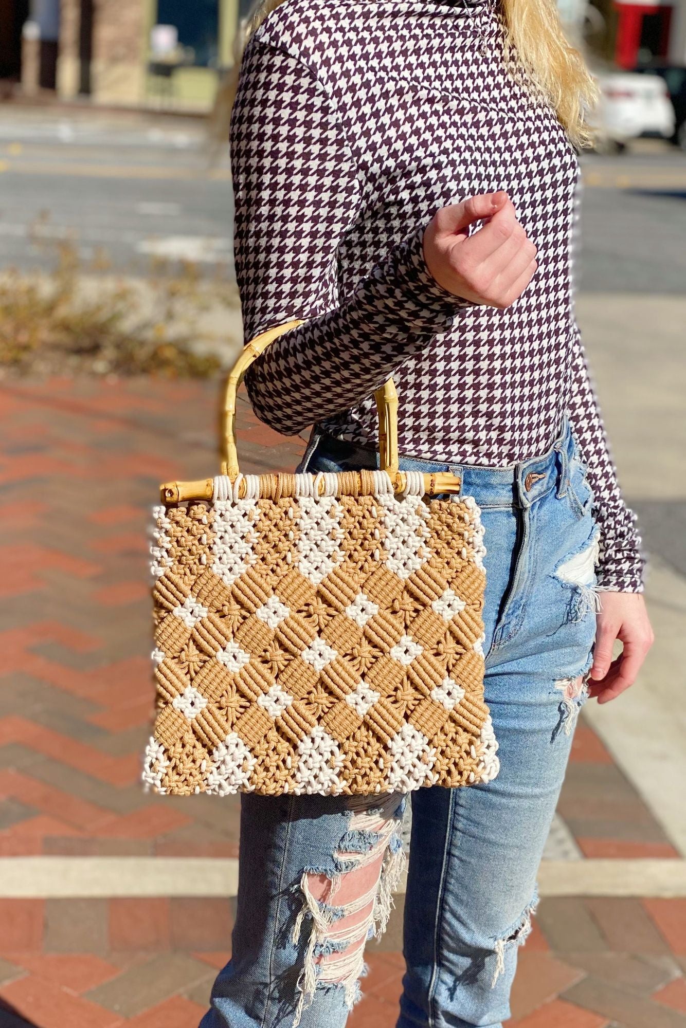 Stylish handwoven tan and white macrame handbag held by a woman wearing a patterned long-sleeve shirt and distressed jeans, with a vibrant outdoor background.