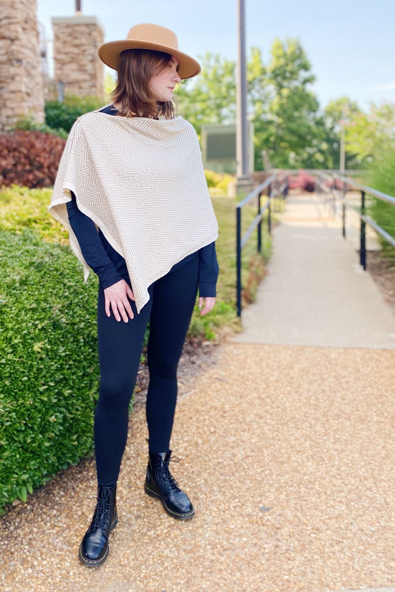 Woman wearing a stylish beige herringbone-patterned poncho over a black long-sleeve shirt and black leggings, paired with black lace-up combat boots, standing on a gravel path surrounded by greenery.