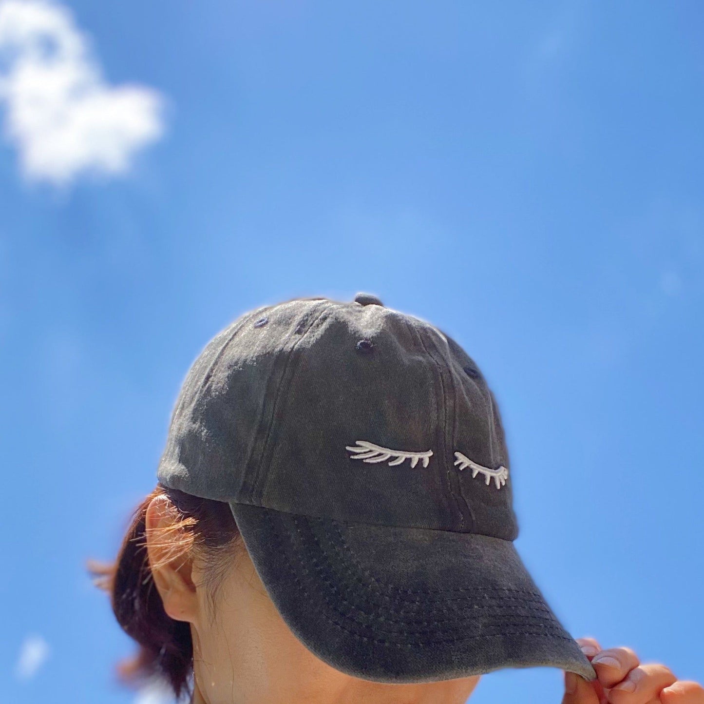 Black baseball cap with white eyelash embroidery worn by a woman against a bright blue sky.