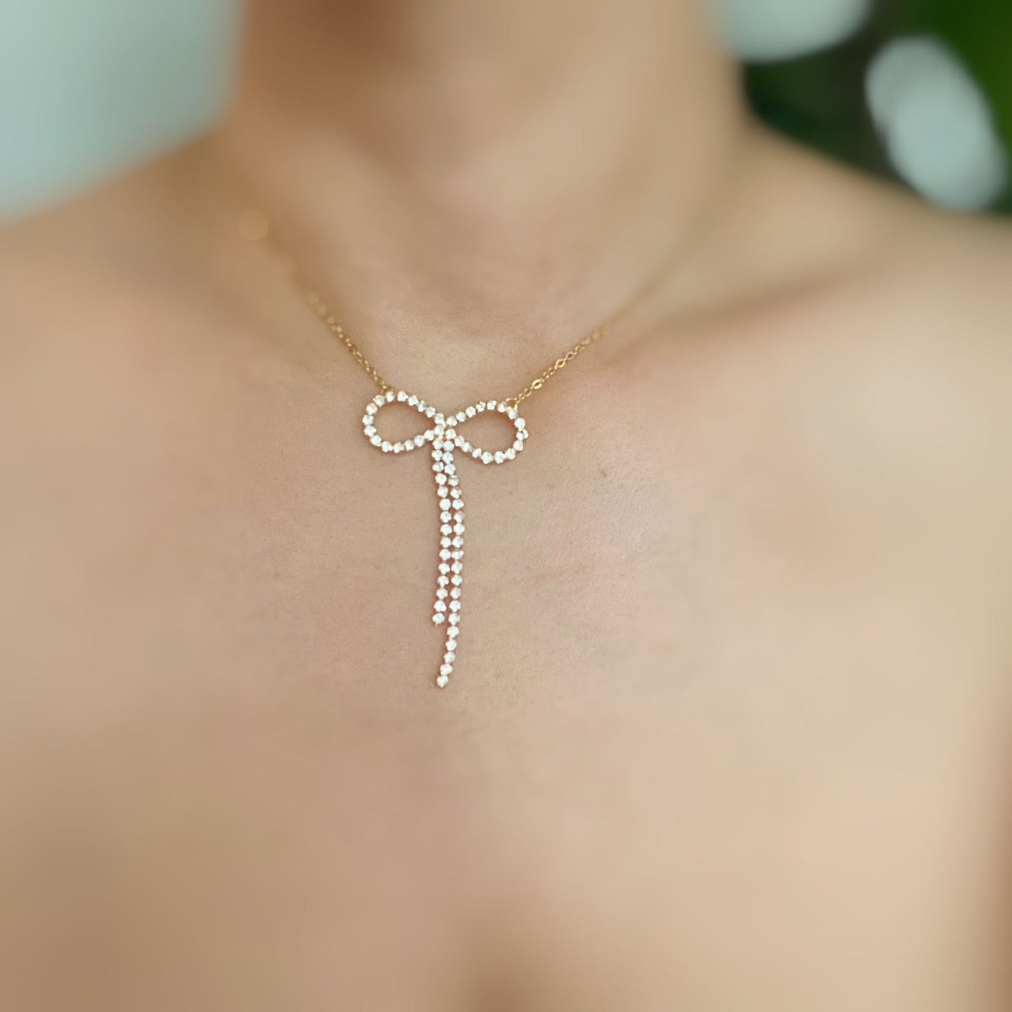 Elegant gold necklace featuring a crystal-studded bow pendant with cascading strands, displayed on a bare neckline against a soft, blurred background.