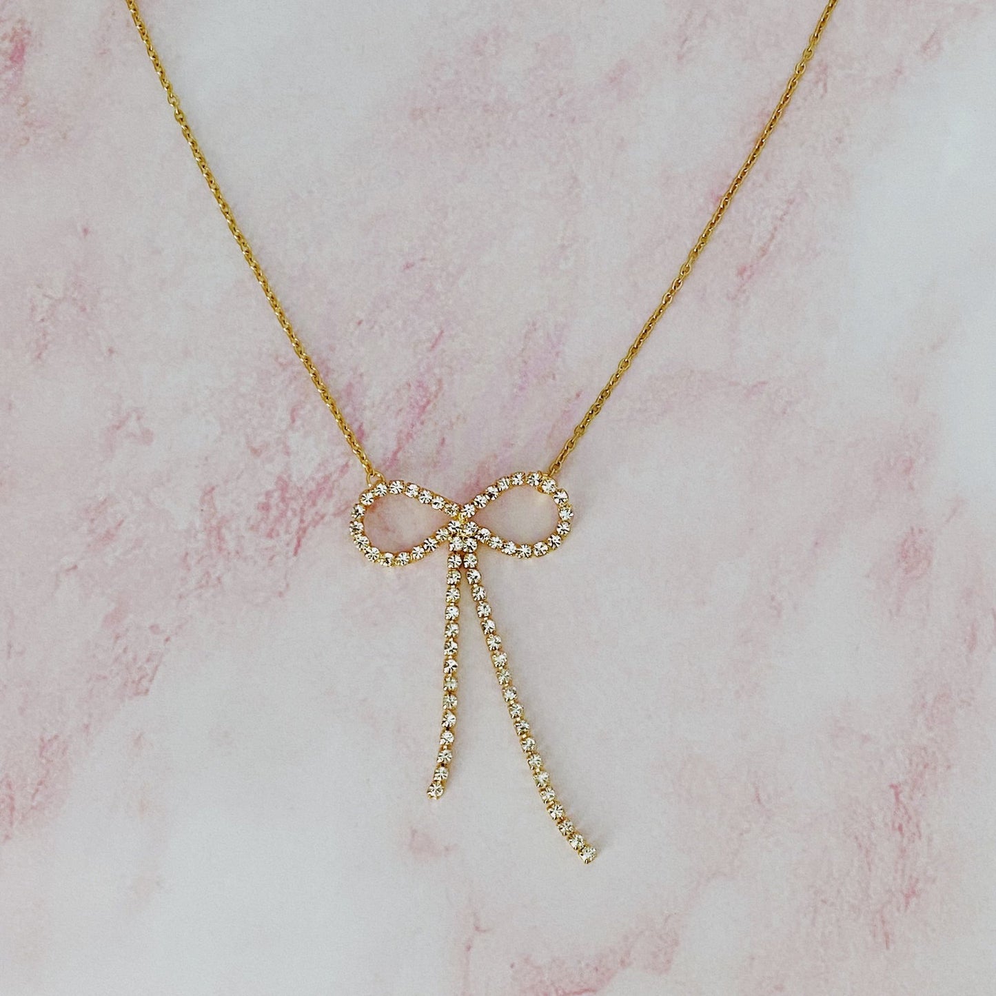 Gold necklace featuring a diamond-studded bow pendant on a delicate chain, set against a soft pink marble background.