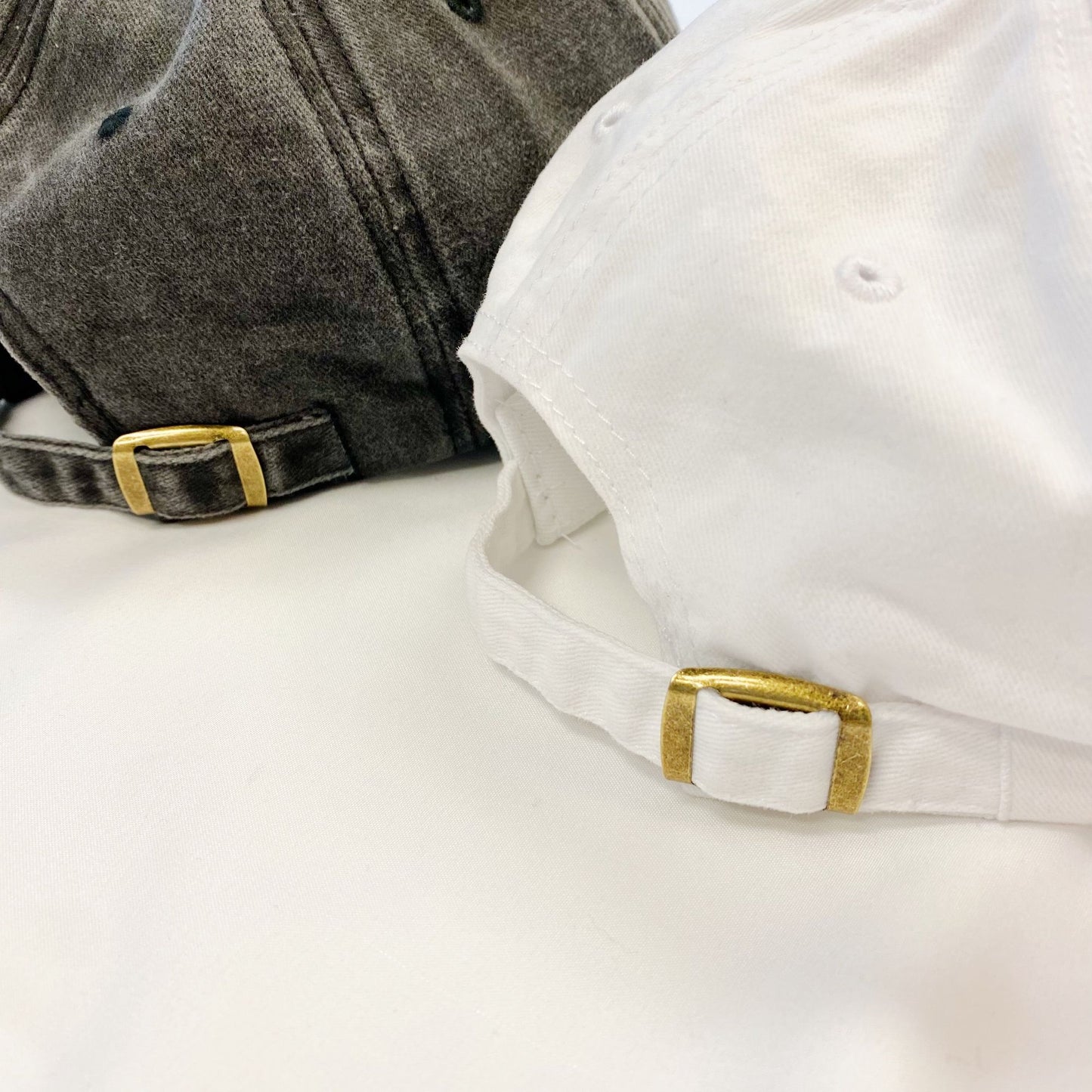 Close-up of a gray and a white baseball cap, showcasing their adjustable straps with gold metal buckles against a neutral background.