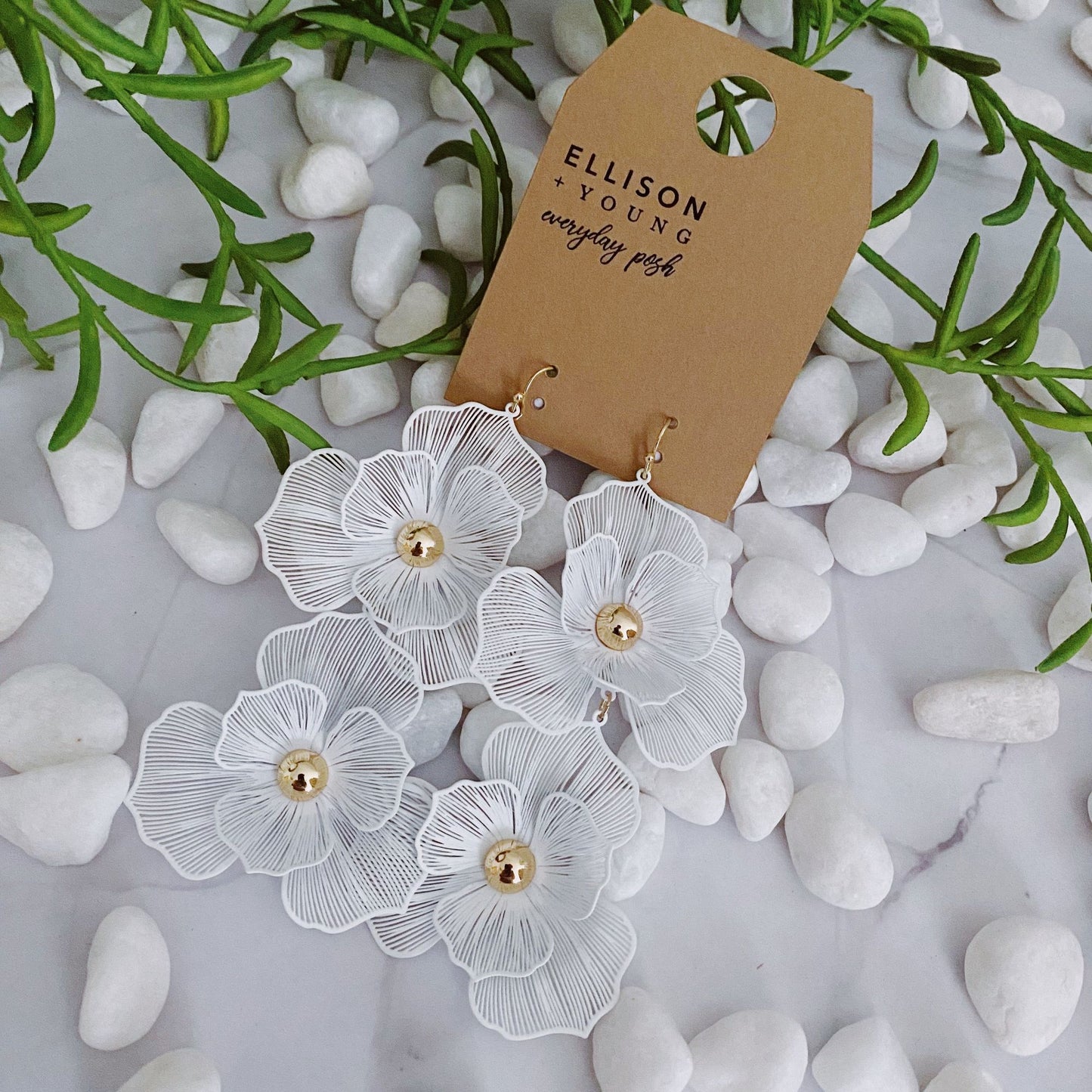 Elegant white floral earrings with gold accents displayed on a natural stone background, featuring a brown tag that reads "ELLISON YOUNG everyday posh."