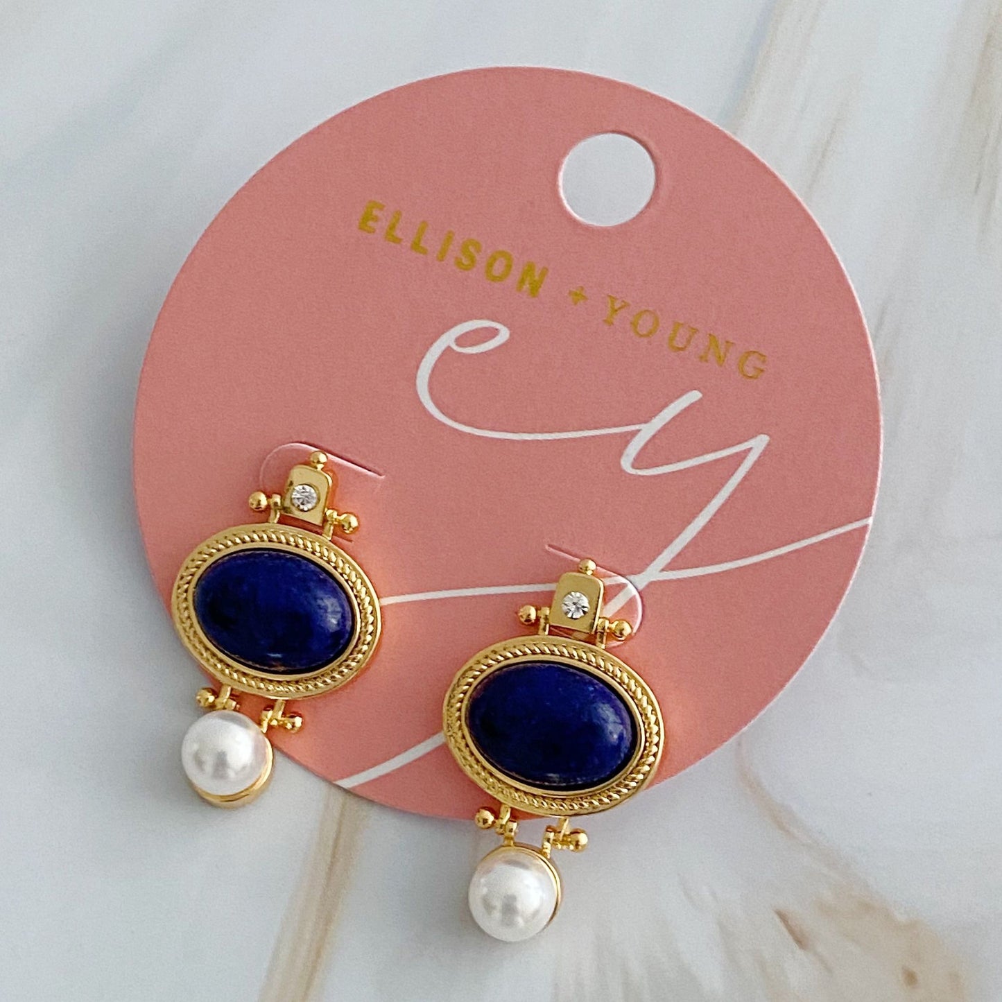 Elegant gold earrings featuring deep blue lapis lazuli stones, accented with sparkling clear crystals and white pearl drops, displayed on a round pink card.