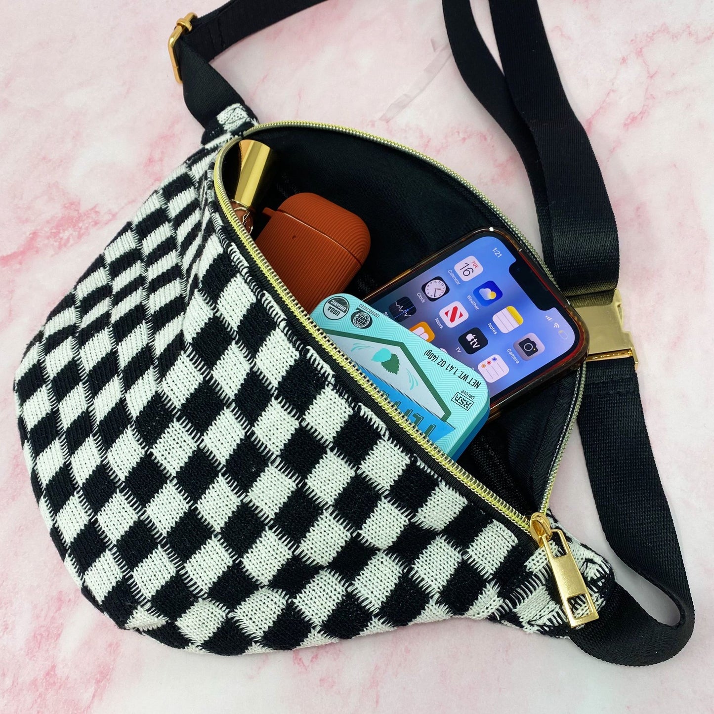 Checkered fanny pack with gold zipper, filled with a smartphone, small speaker, and cash, displayed against a pink marble background.