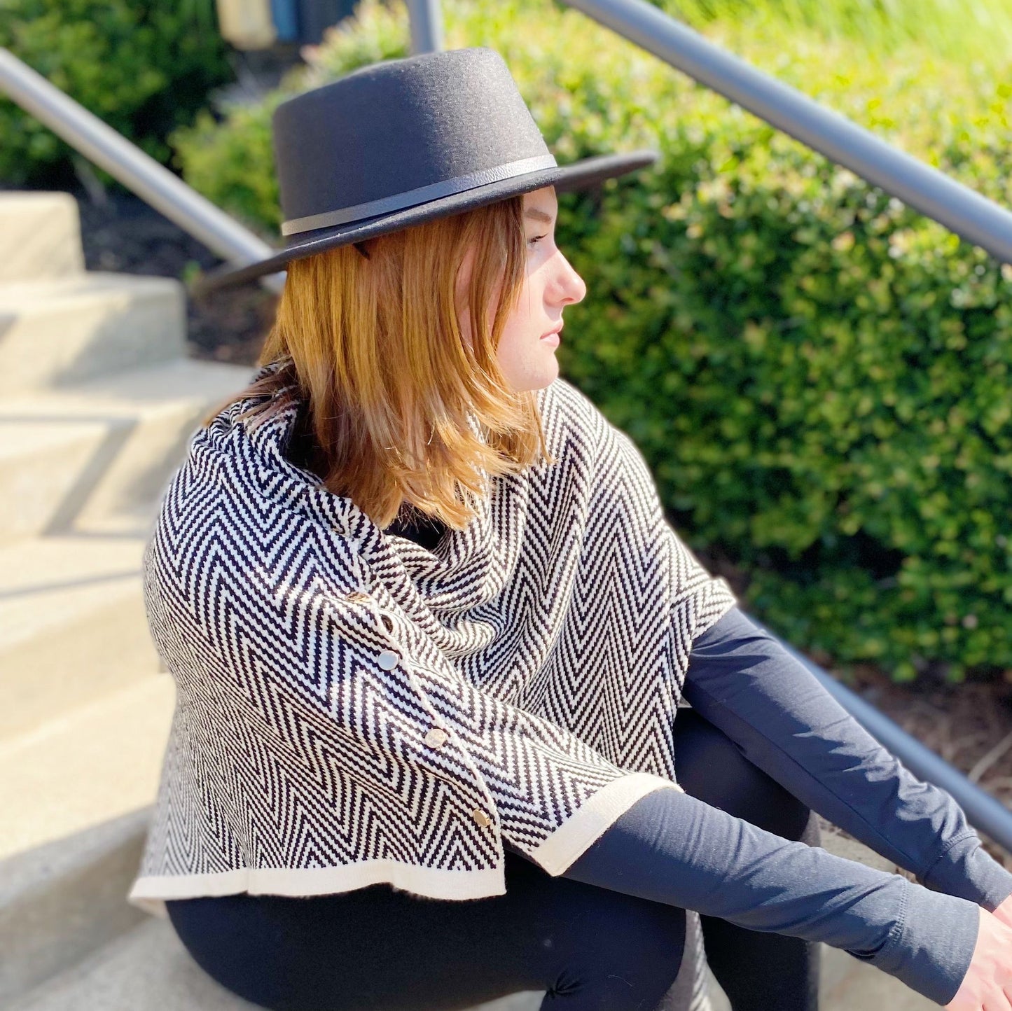 Stylish woman wearing a black and white herringbone poncho and a wide-brimmed black hat, sitting on outdoor stairs with lush green bushes in the background.