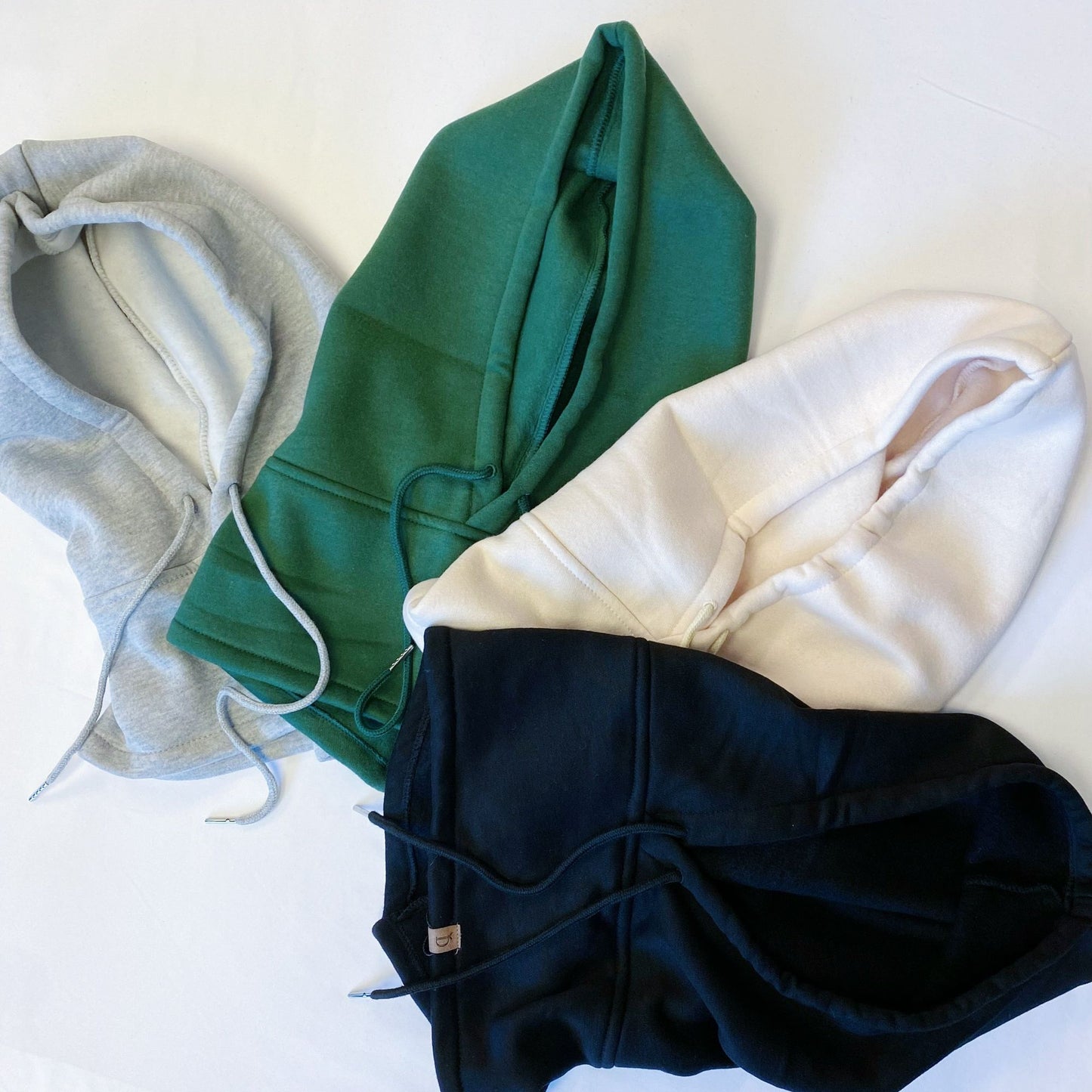 Four stylish hoodies in gray, green, cream, and black laid flat on a white background, showcasing their drawstrings and soft fabric.