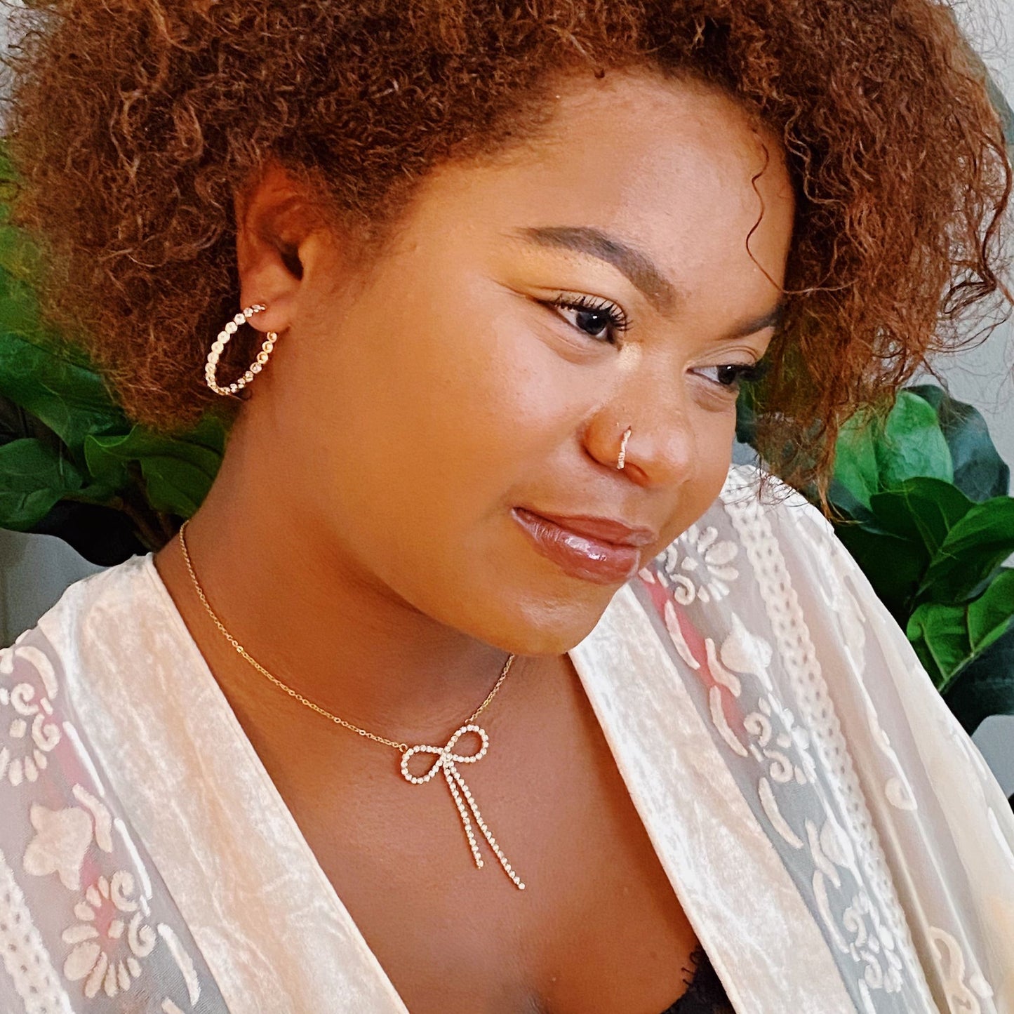 Elegant gold necklace featuring a bow design and sparkling rhinestone accents, paired with stylish hoop earrings, set against a lush green background.