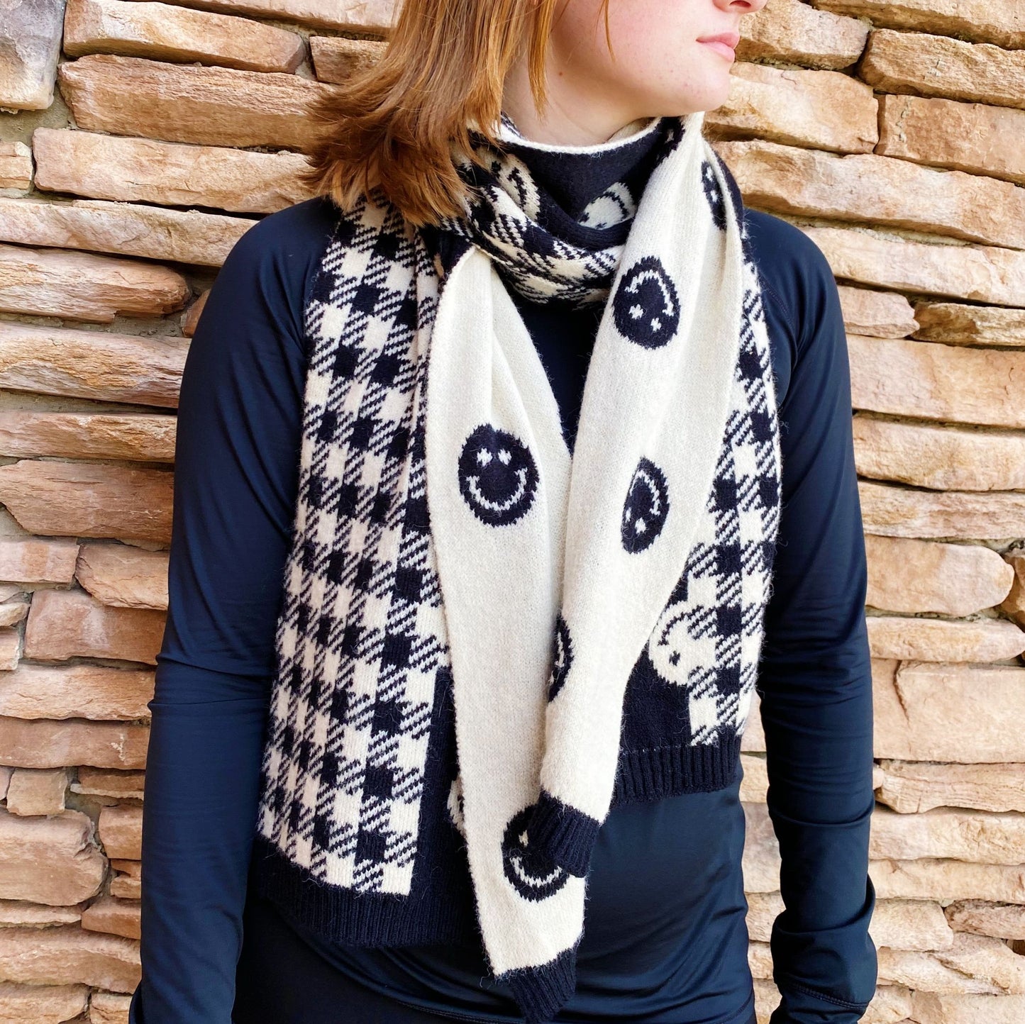Stylish black and white patterned scarf featuring smiley faces, worn by a woman against a stone wall backdrop, perfect for adding a cheerful touch to any outfit.