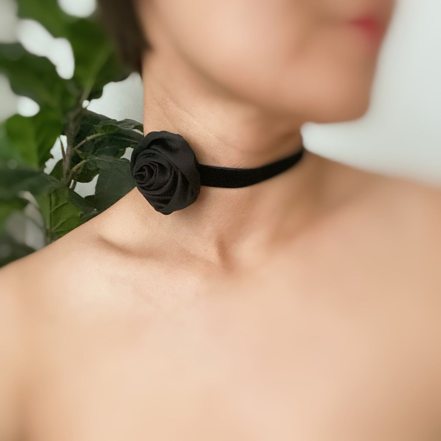 Close-up of a woman wearing a chic black velvet choker with a rose detail, showcasing a stylish and modern accessory against a softly blurred background of green leaves.