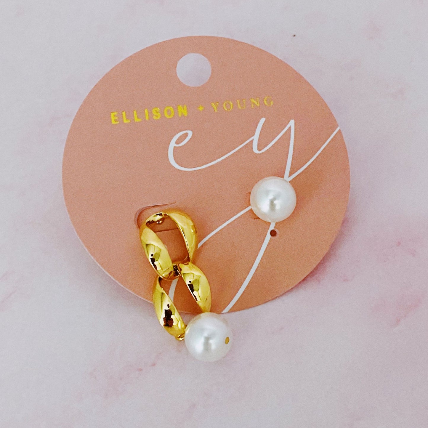 Gold chain and pearl stud earrings on a pink display card by Ellison + Young.
