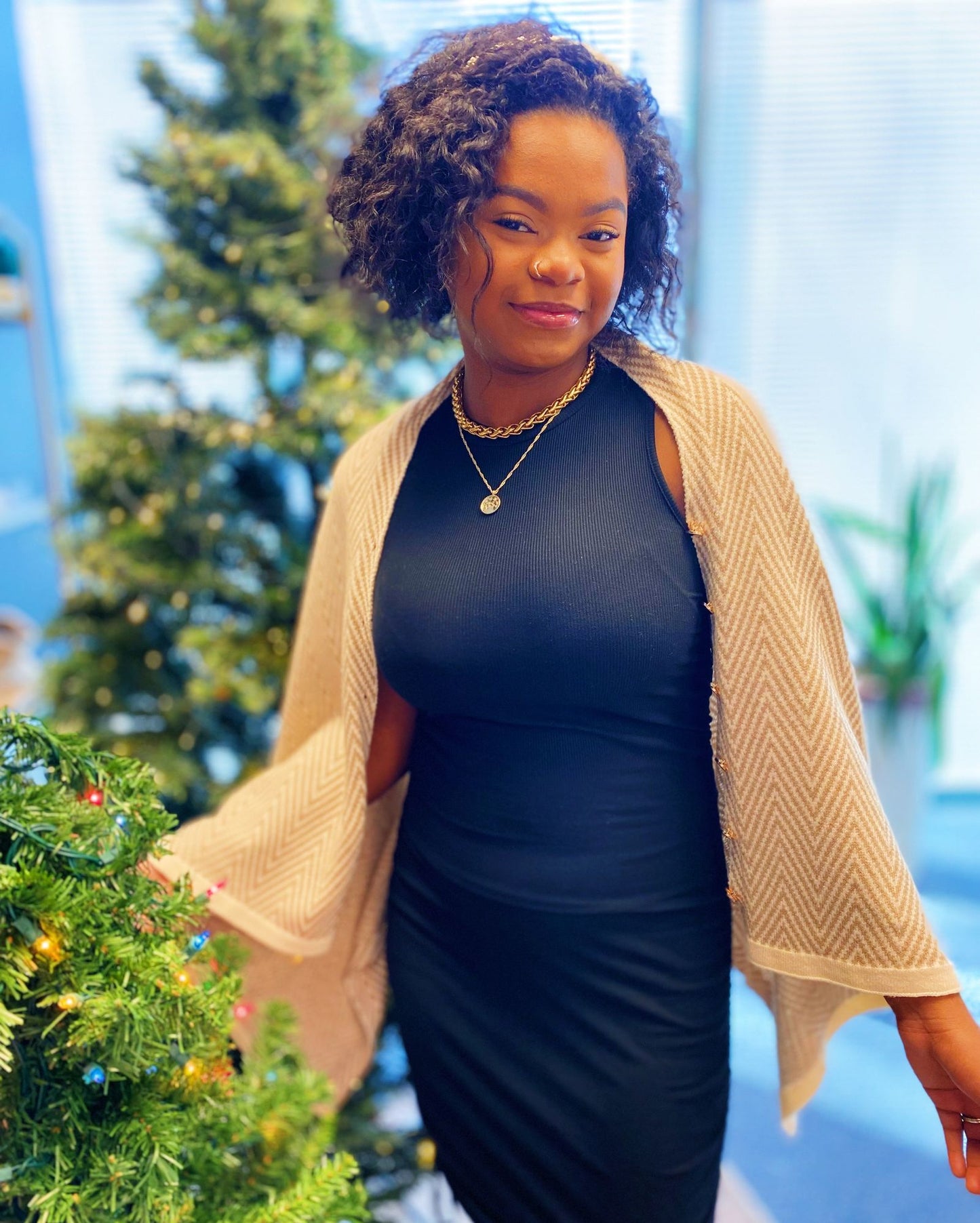 A woman in a stylish black dress and cream-colored cardigan stands beside a decorated Christmas tree, showcasing her chic holiday outfit.