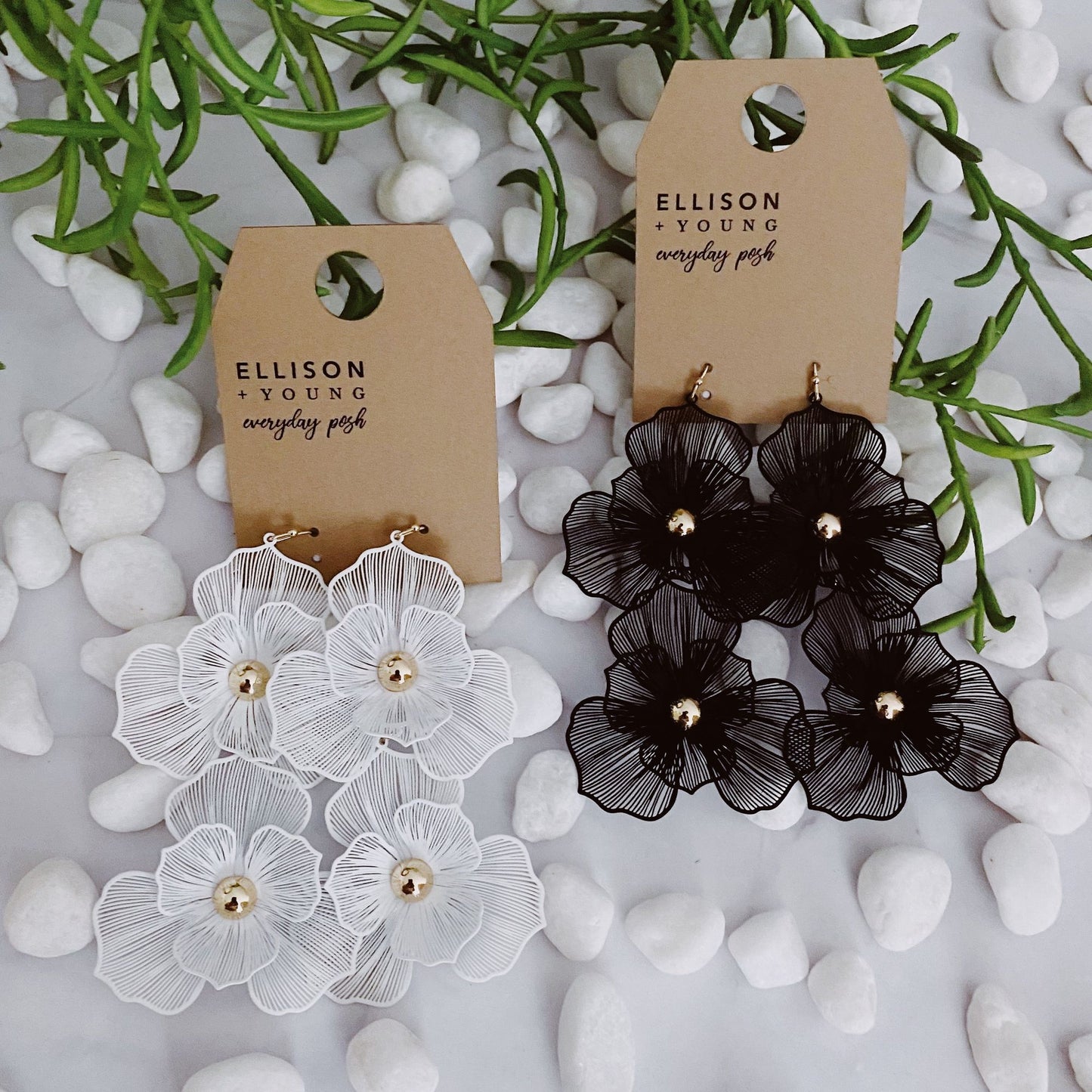 Elegant floral earrings in white and black, featuring mesh flower designs with gold accents, displayed on eco-friendly tags against a backdrop of smooth white stones and greenery.