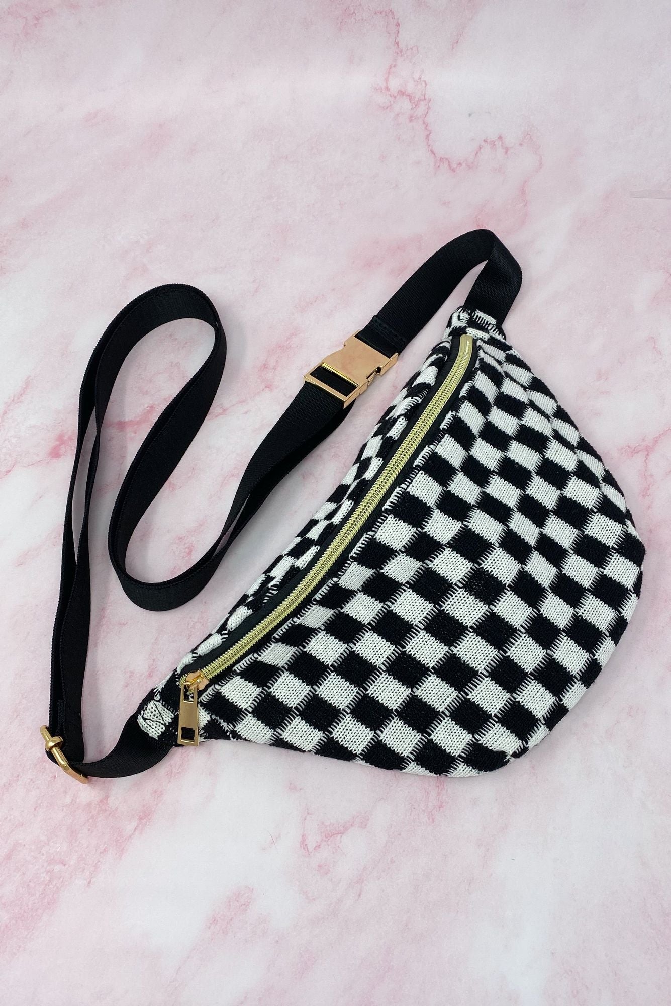 Black and white checkered fanny pack with a gold zipper and adjustable strap, set against a soft pink background.