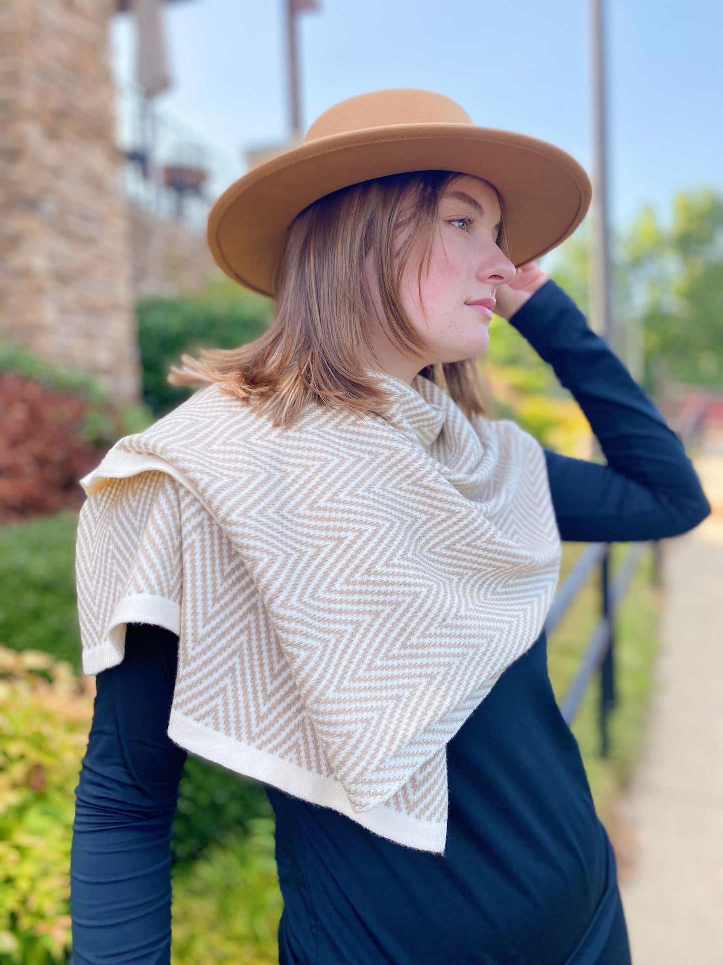 A woman wearing a stylish tan hat and a patterned beige and white scarf, posing outdoors against a background of greenery and a stone wall.
