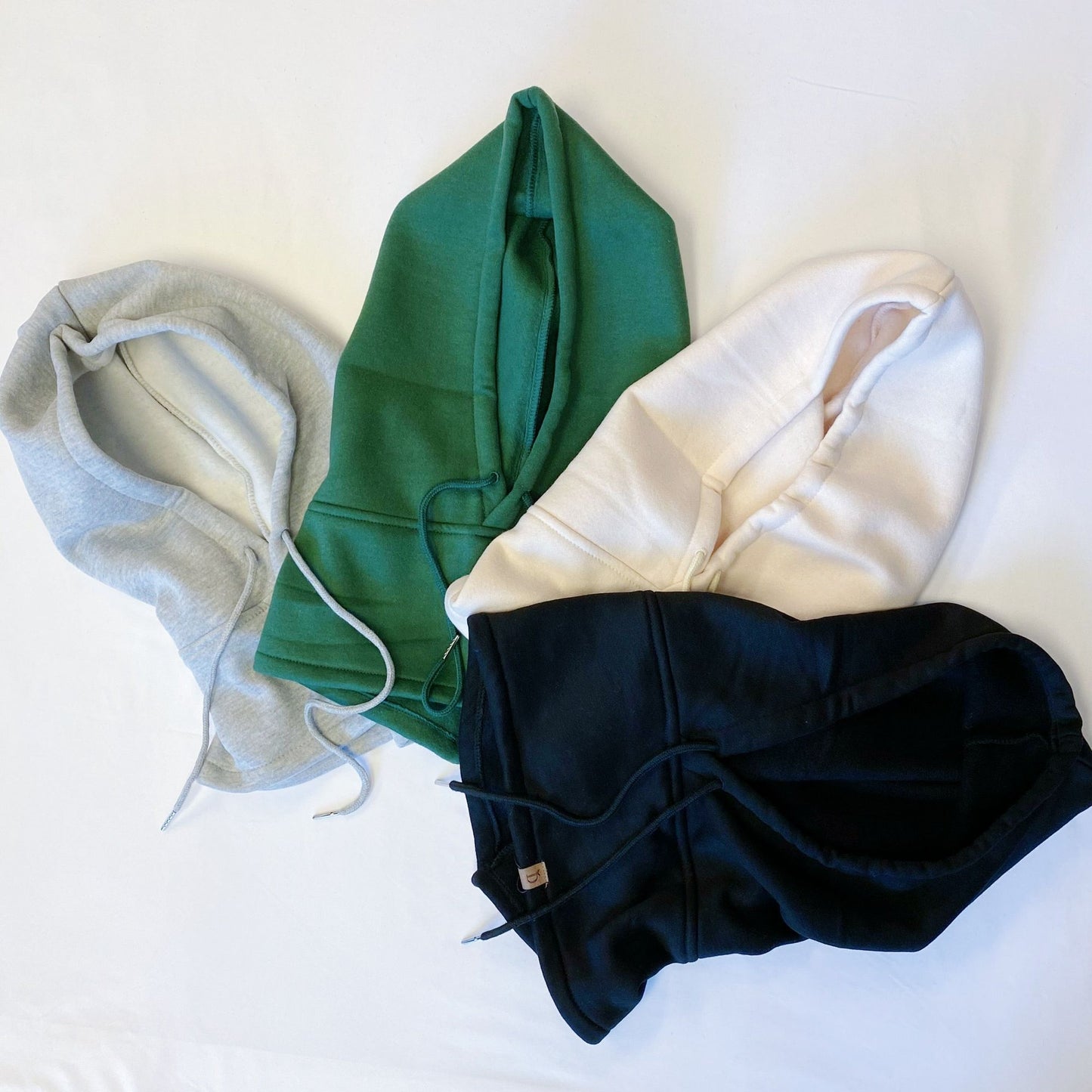 Three stylish hoodies in gray, green, and cream resting on a white surface, showcasing their cozy design and drawstring details.