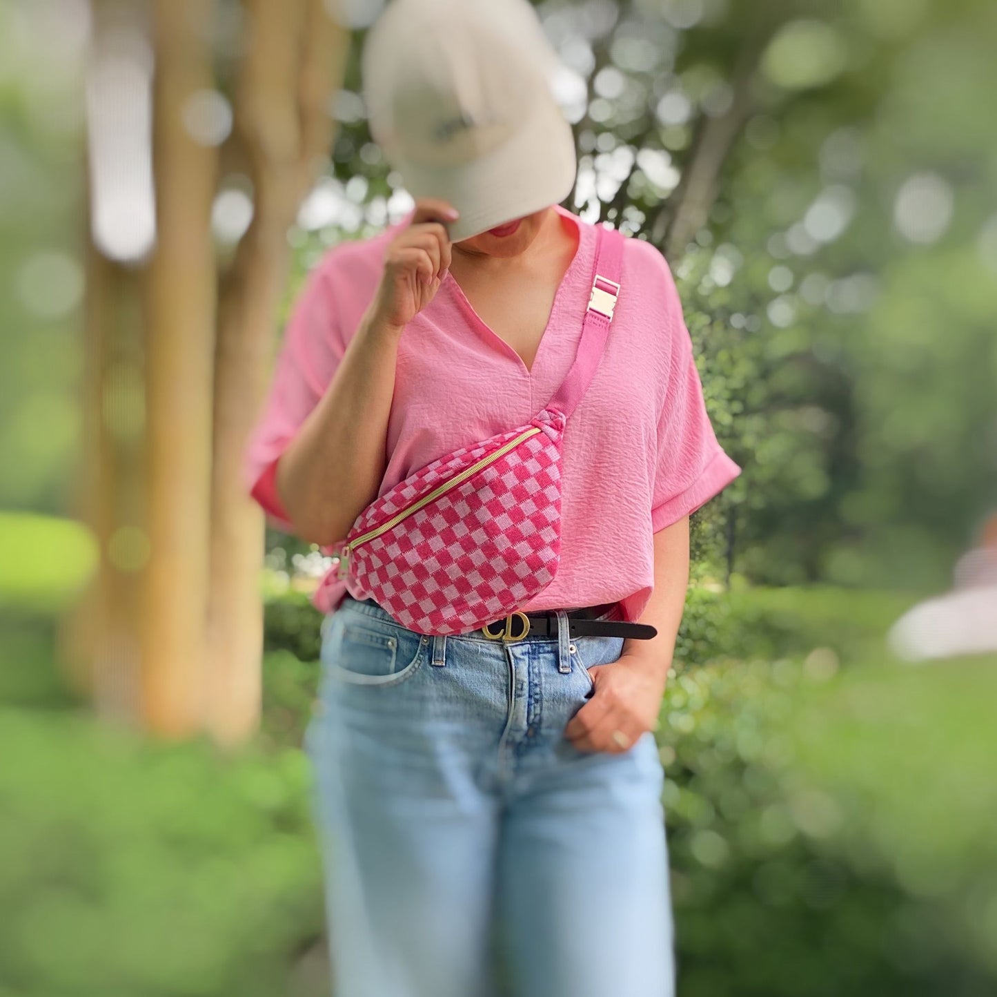 Stylish woman wearing a pink v-neck shirt and blue jeans, posing outdoors while holding a pink checkered fanny pack, with a beige cap partially covering her face.