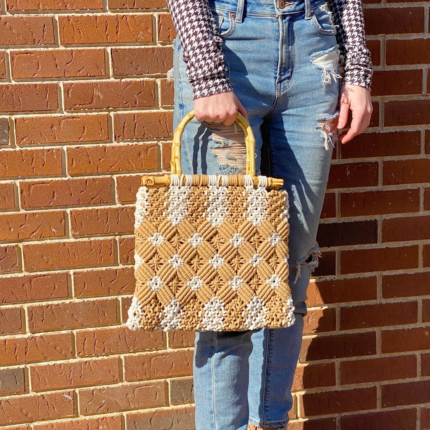 Stylish handwoven straw handbag with bamboo handle, featuring a geometric pattern in brown and cream, held by a model wearing distressed blue jeans against a brick wall.