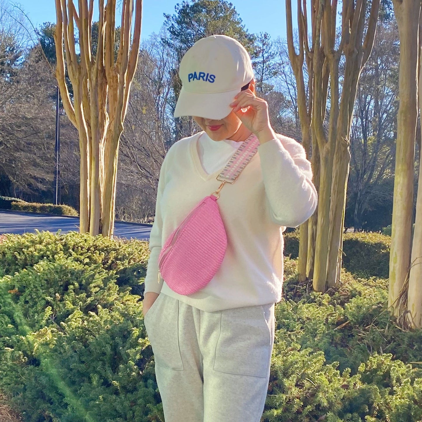 A person wearing a cozy white sweater and gray sweatpants, standing outdoors with tall trees in the background, holding a stylish pink crossbody bag.