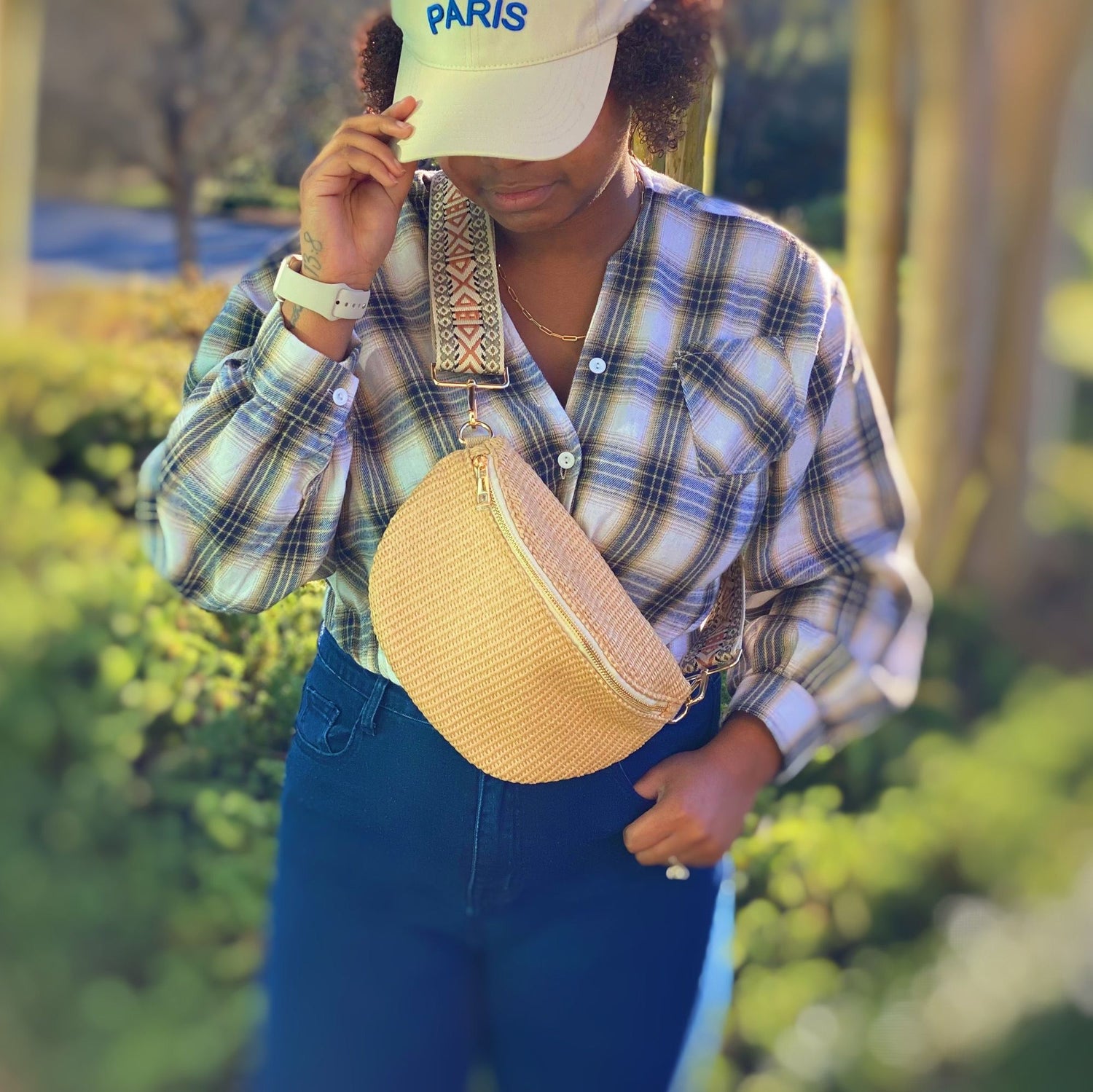 Stylish woman wearing a plaid shirt and high-waisted jeans, holding a beige crossbody bag with a decorative strap, set against a lush green background.