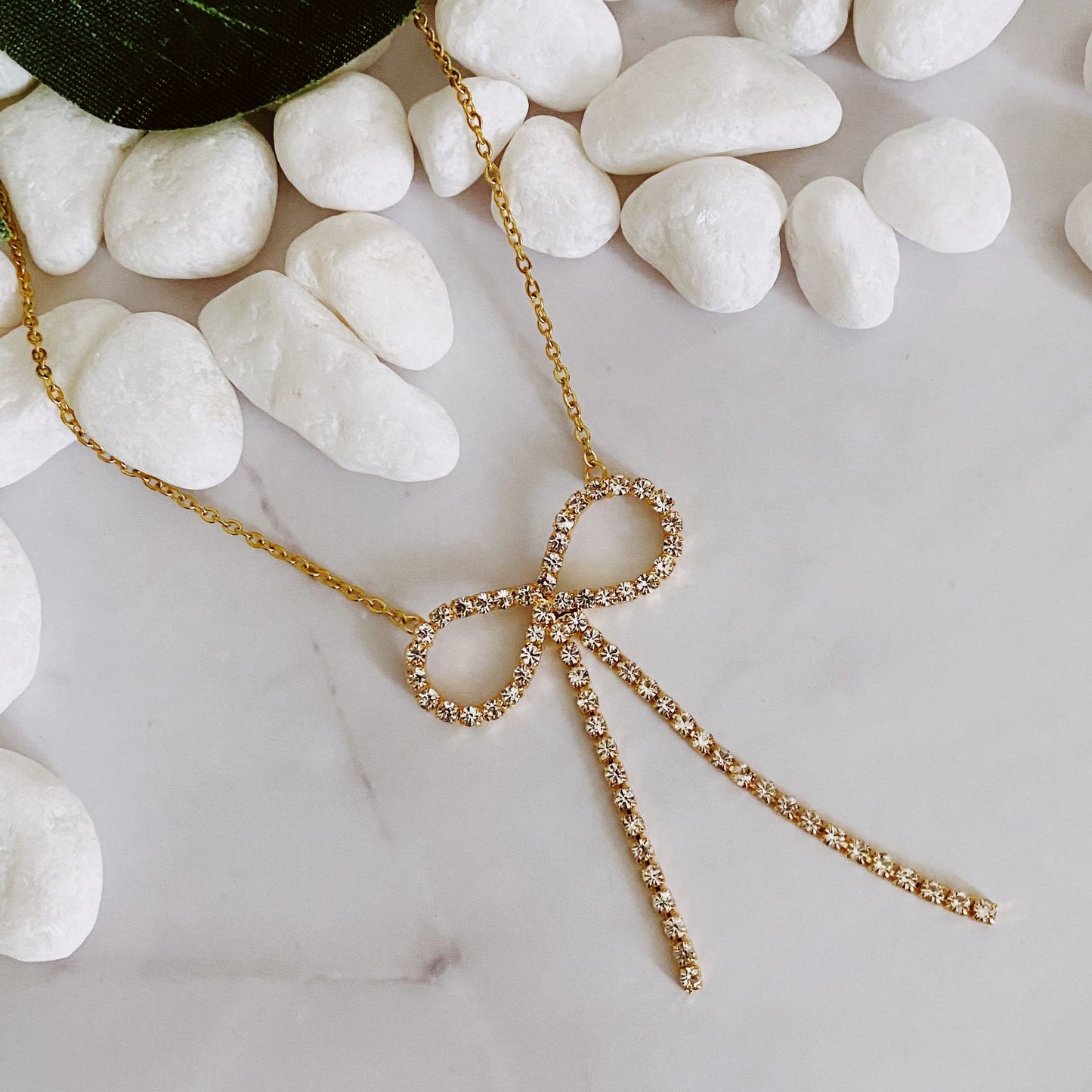 Gold necklace with a sparkling rhinestone bow pendant resting on white pebbles against a marble background.