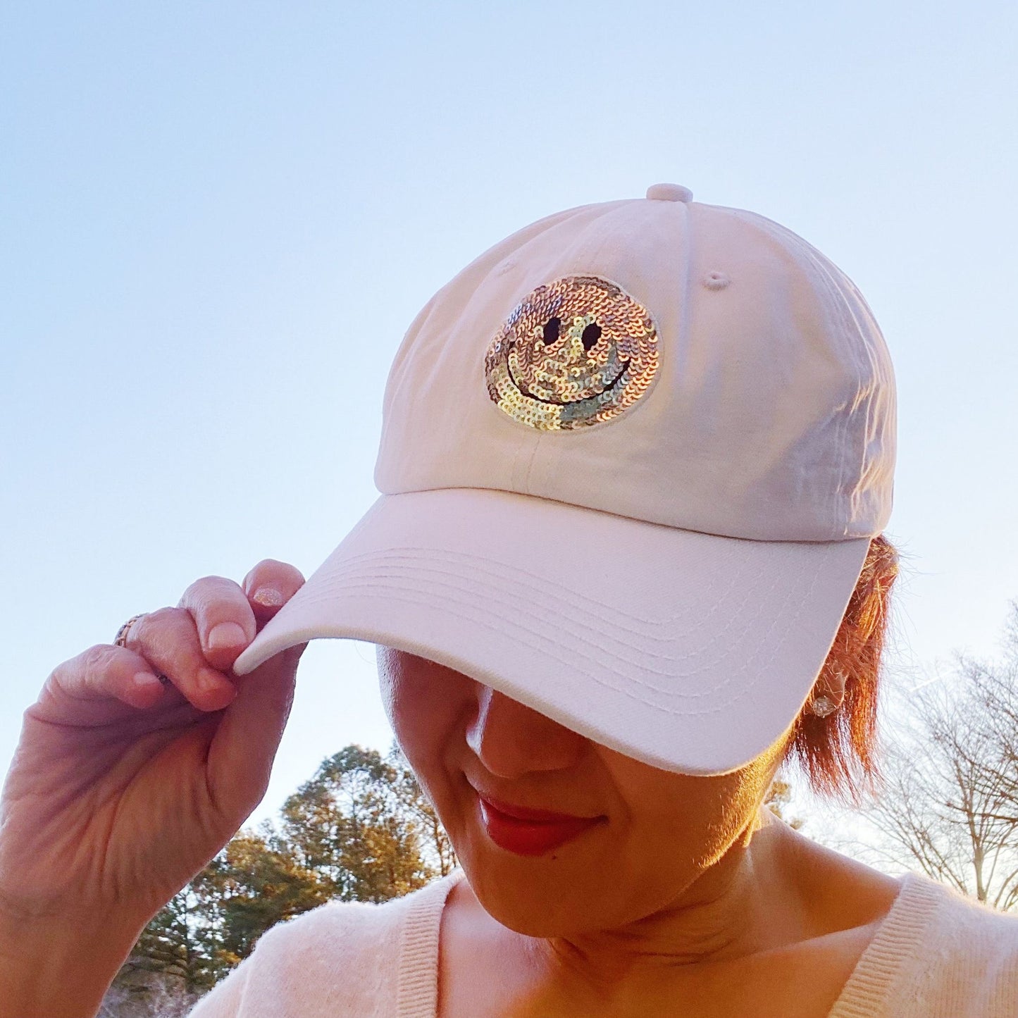 Light beige cap with a sparkling sequin smiley face emblem, held casually against a soft outdoor background, perfect for sunny days and enhancing your laid-back style.