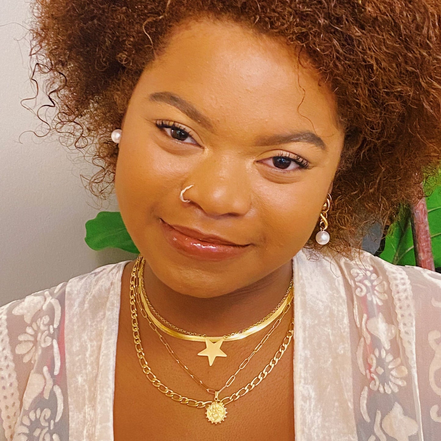 Layered gold necklaces featuring a star and sun pendant, worn with a light, sheer white top, against a backdrop of greenery.