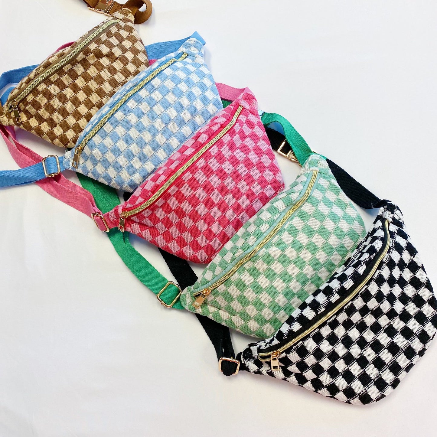 Colorful checkered fanny packs in pink, blue, green, brown, and black arranged in a row, featuring gold zippers and adjustable straps on a white background.