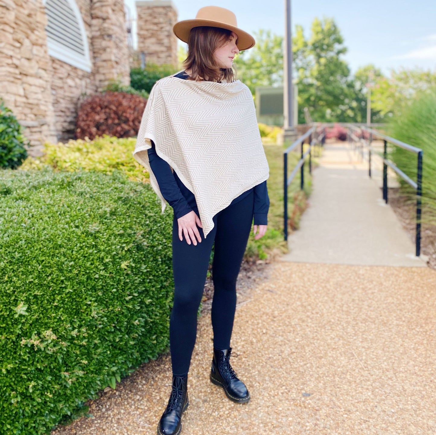 Stylish woman wearing a beige chevron-patterned poncho over a black long-sleeve top and black leggings, complemented by lace-up black combat boots, standing outdoors near lush greenery and a gravel path.