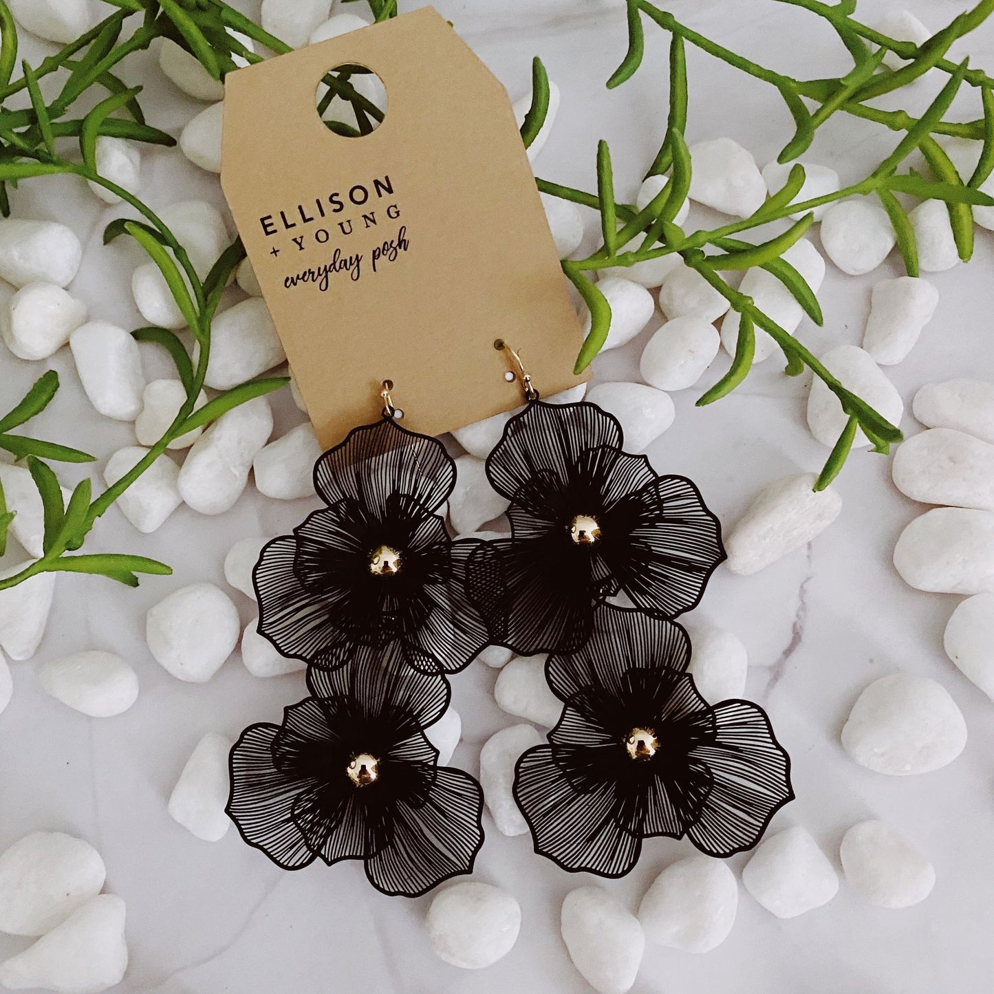 Elegant black flower earrings displayed on a brown tag surrounded by white stones and greenery. Perfect accessory for a chic look.