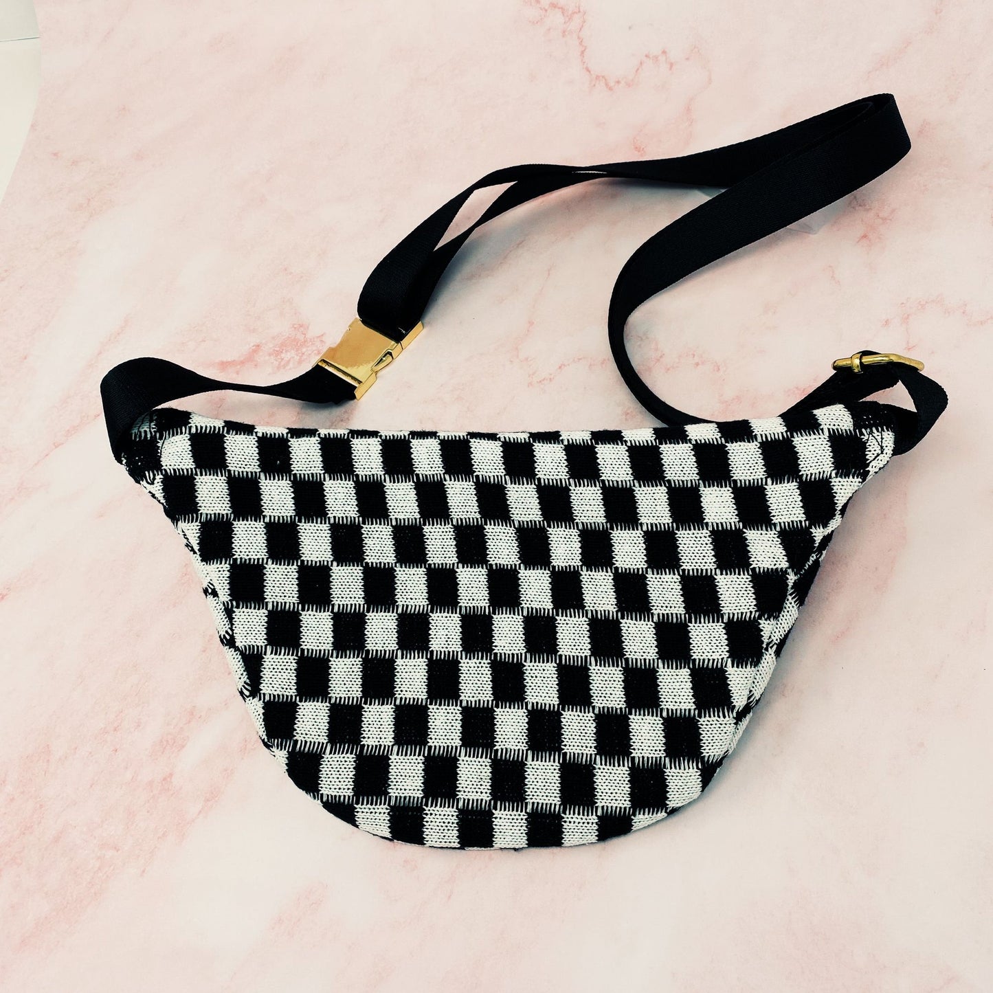 Black and white checkered fanny pack with adjustable black strap and gold buckle, placed on a pink marble background.