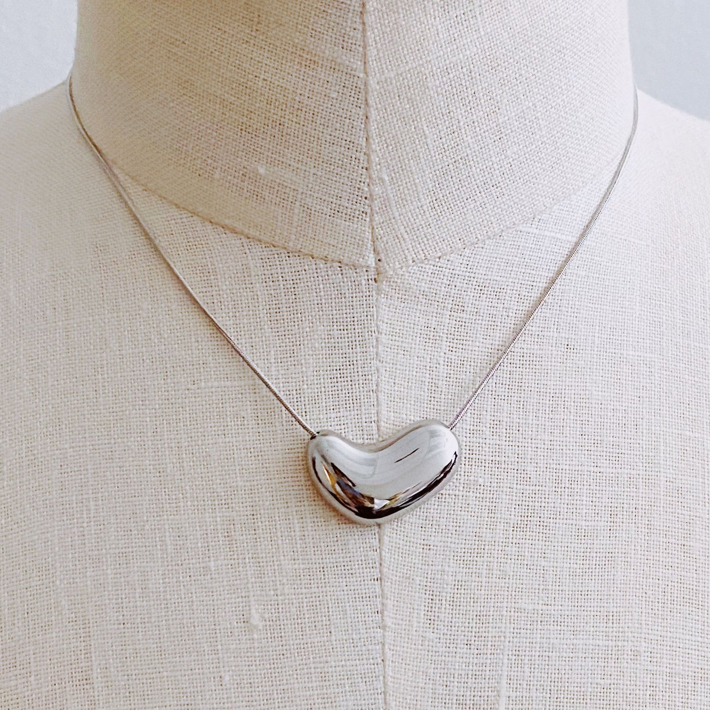 Stylish polished silver heart-shaped pendant necklace displayed on a linen mannequin, featuring a delicate silver chain, perfect for elegant occasions or everyday wear.