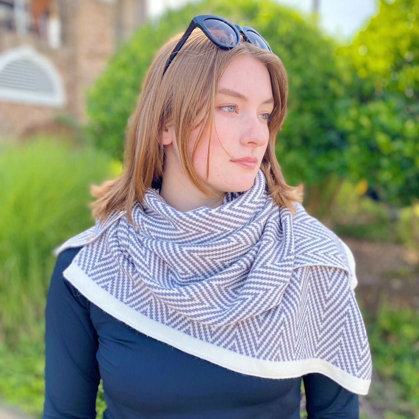 Stylish woman wearing a fashionable herringbone-patterned scarf in neutral colors, paired with a black long-sleeve top, set against a lush green outdoor backdrop.