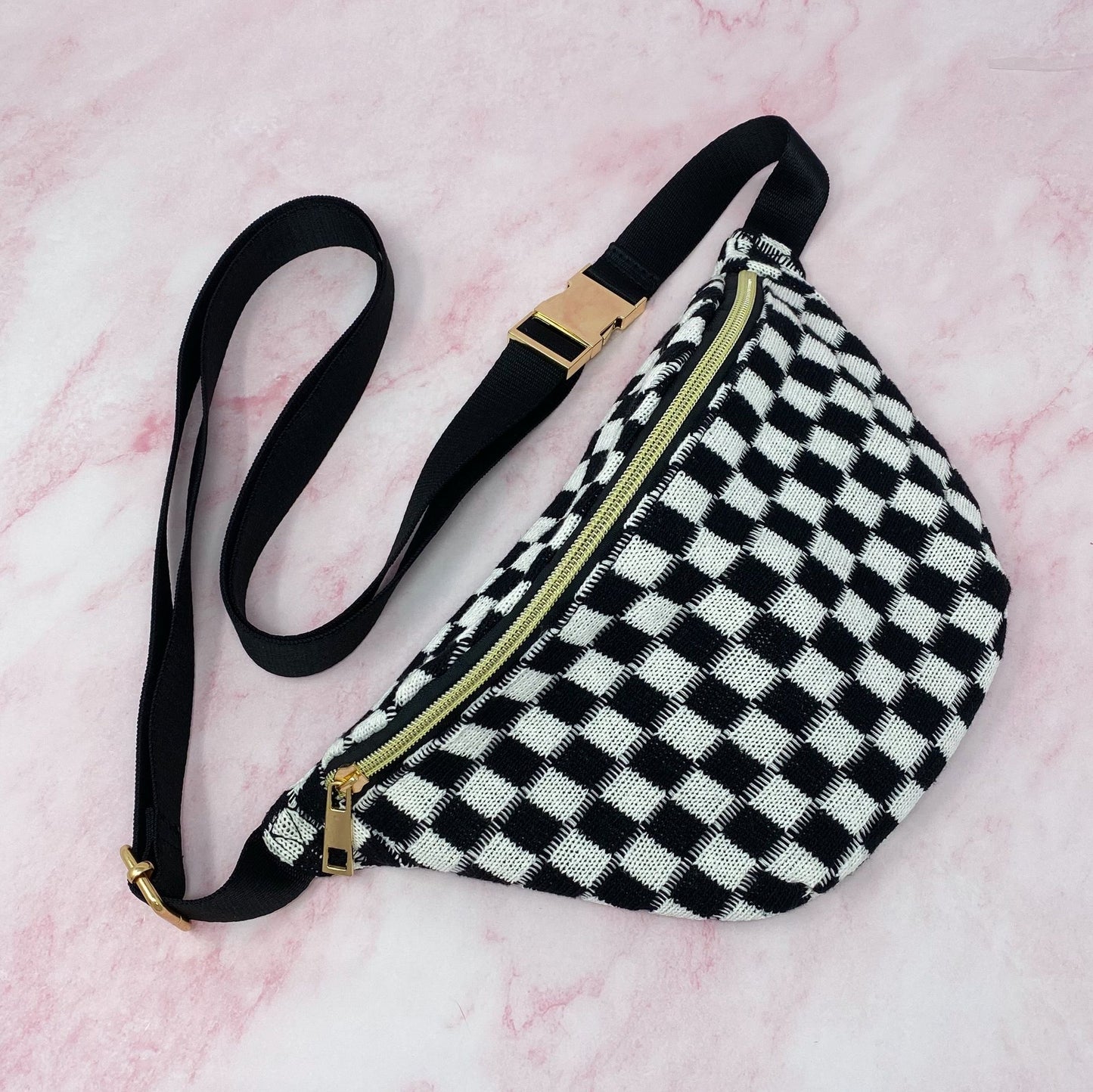 Black and white checkered fanny pack with a gold zipper and adjustable black strap on a pink marble background.