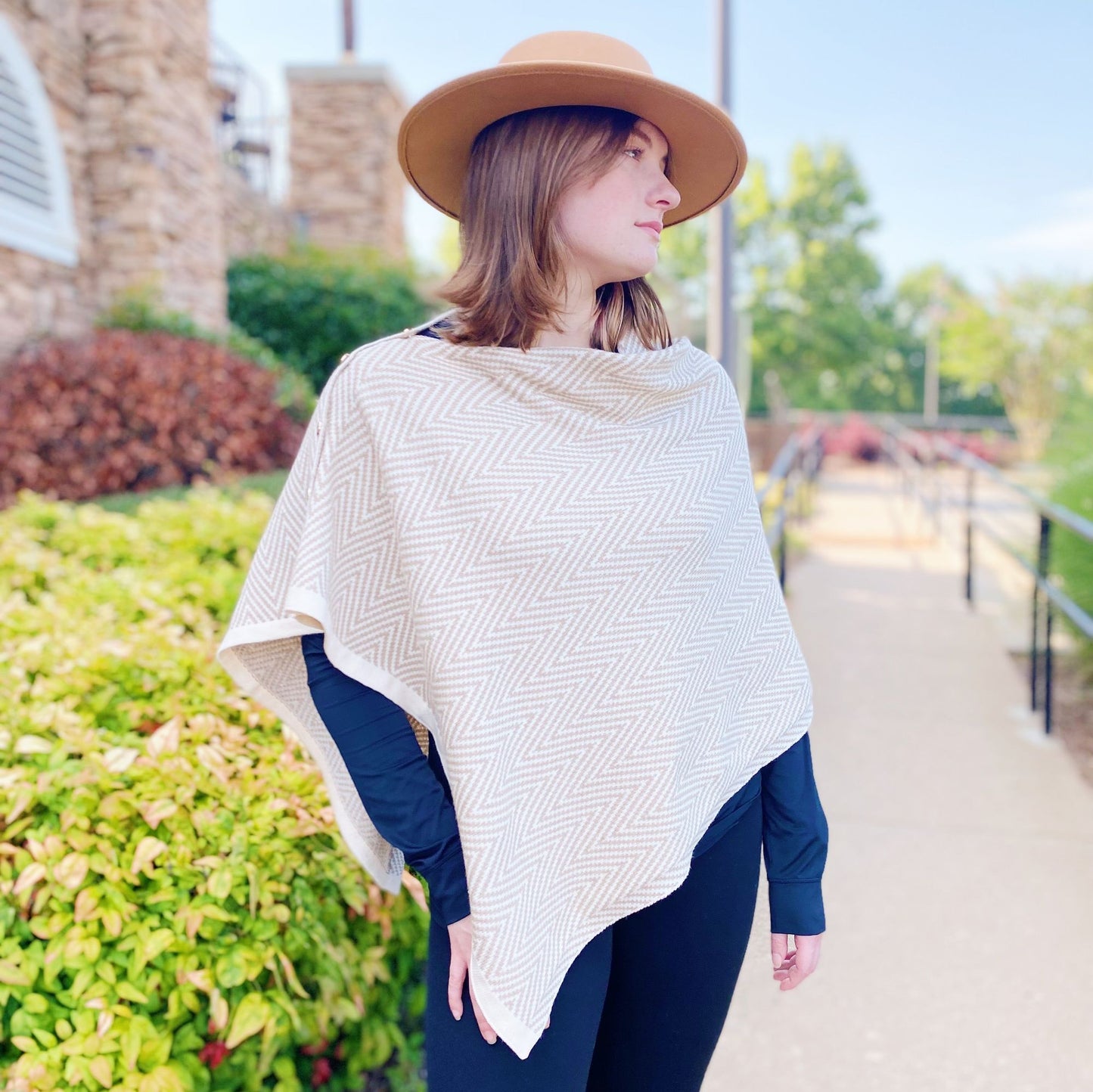 Stylish woman wearing a cream herringbone poncho and a wide-brimmed hat, standing outdoors in a vibrant garden setting with greenery and a pathway.