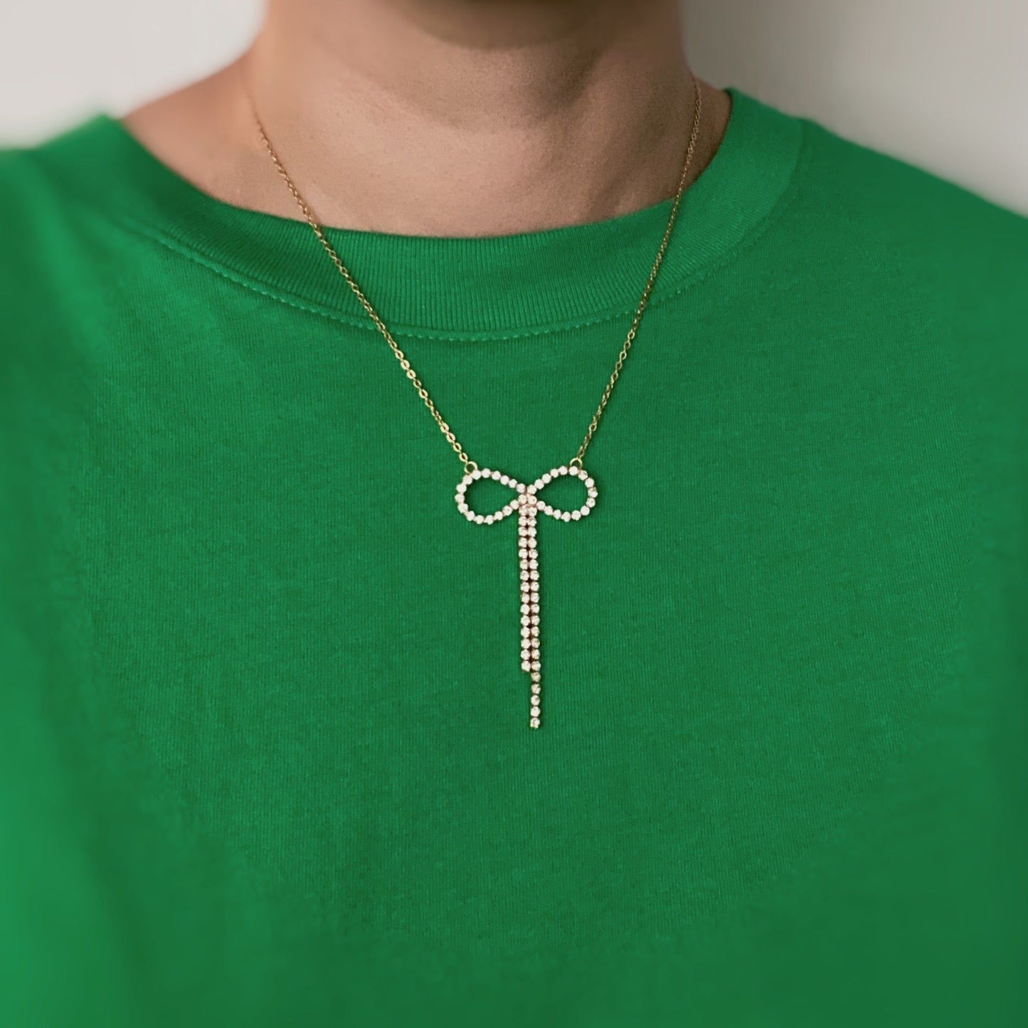 Gold necklace featuring a pearl-encrusted bow pendant, worn over a vibrant green shirt.