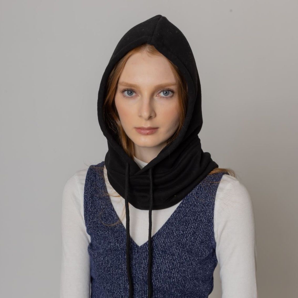 A stylish woman wearing a black hooded scarf over a white long-sleeve top, paired with a blue sleeveless sweater, against a neutral background.