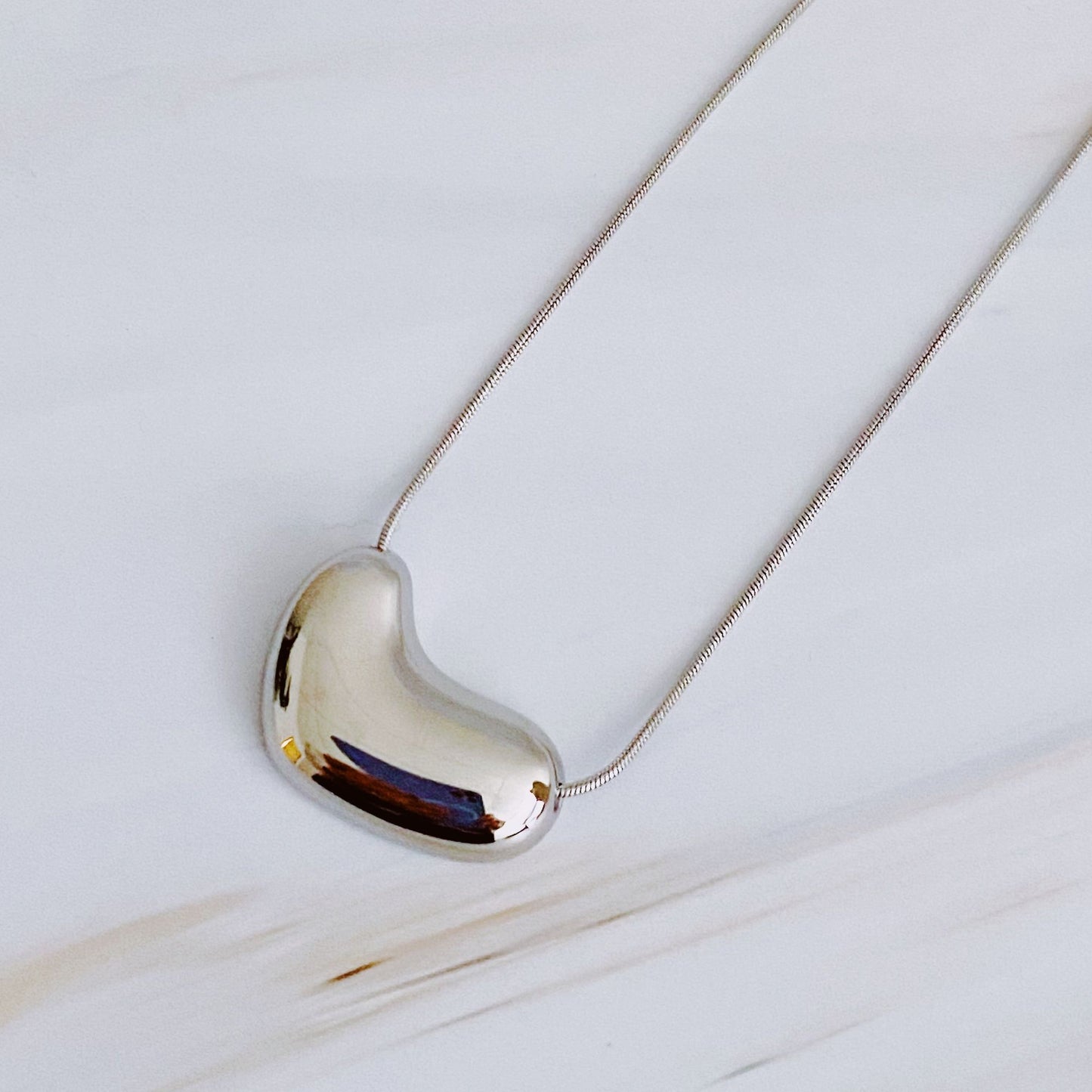 Stylish silver bean-shaped pendant necklace on a delicate chain, displayed on a light marble background.