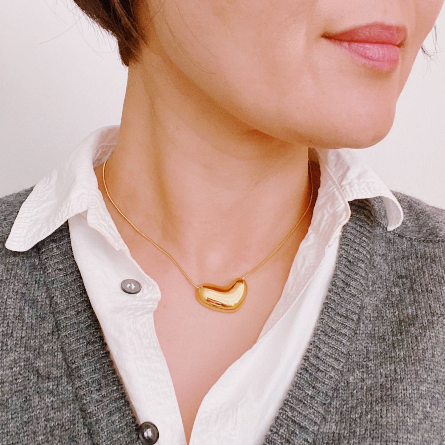 Stylish woman wearing a gold bean-shaped pendant necklace over a white shirt and grey cardigan, showcasing a contemporary accessory for modern fashion lovers.
