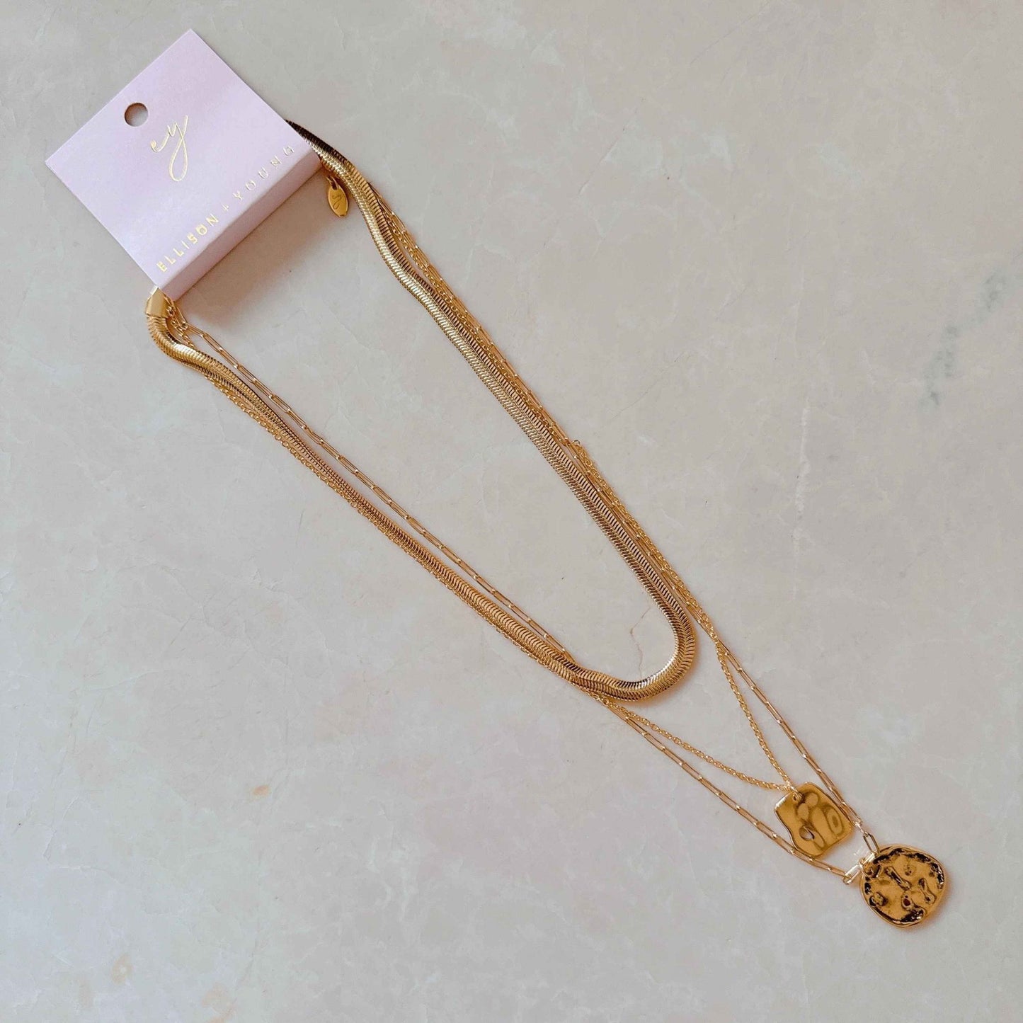 Gold layered necklace with unique charms on a display card, resting on a light-colored textured surface.