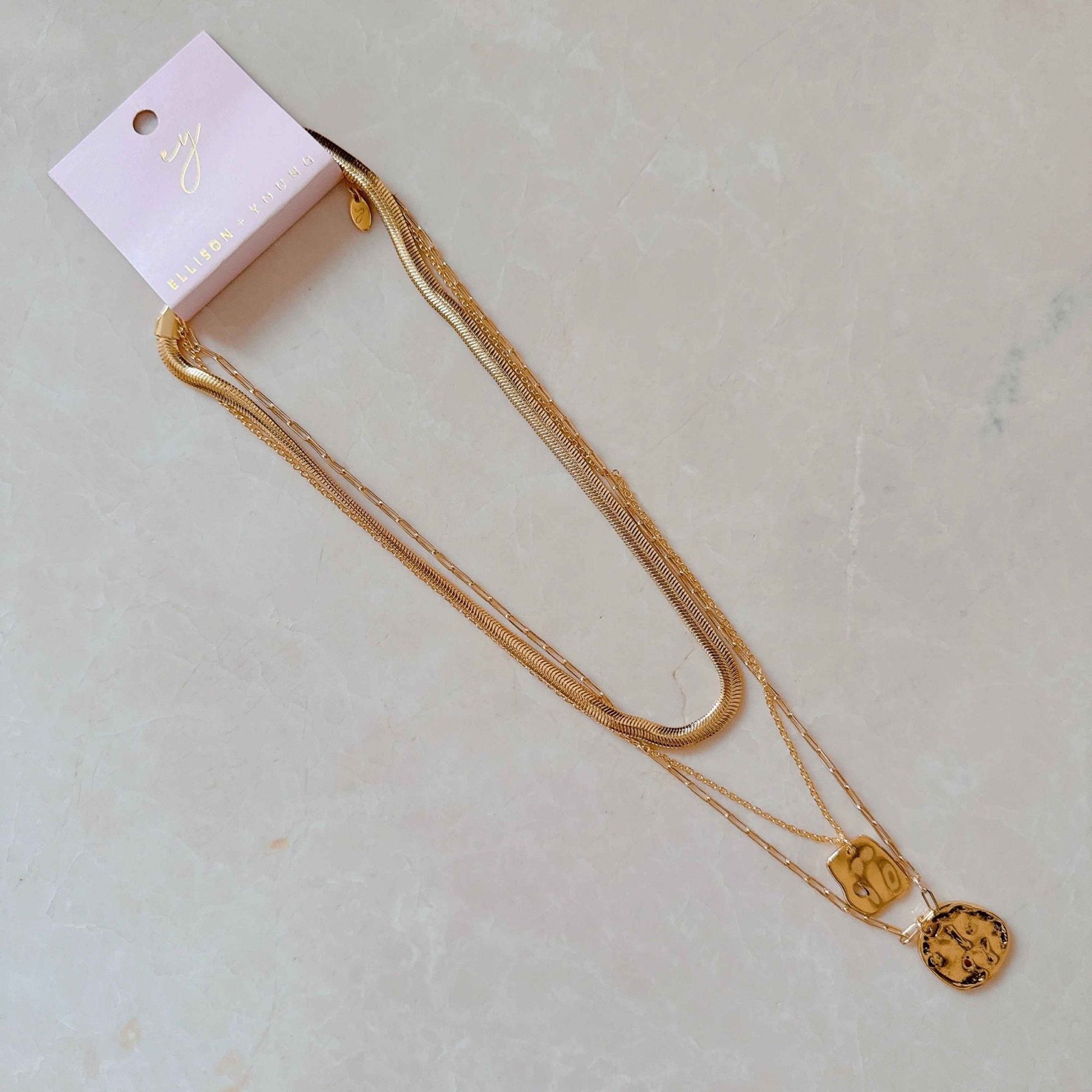 Gold layered necklace with unique charms on a display card, resting on a light-colored textured surface.