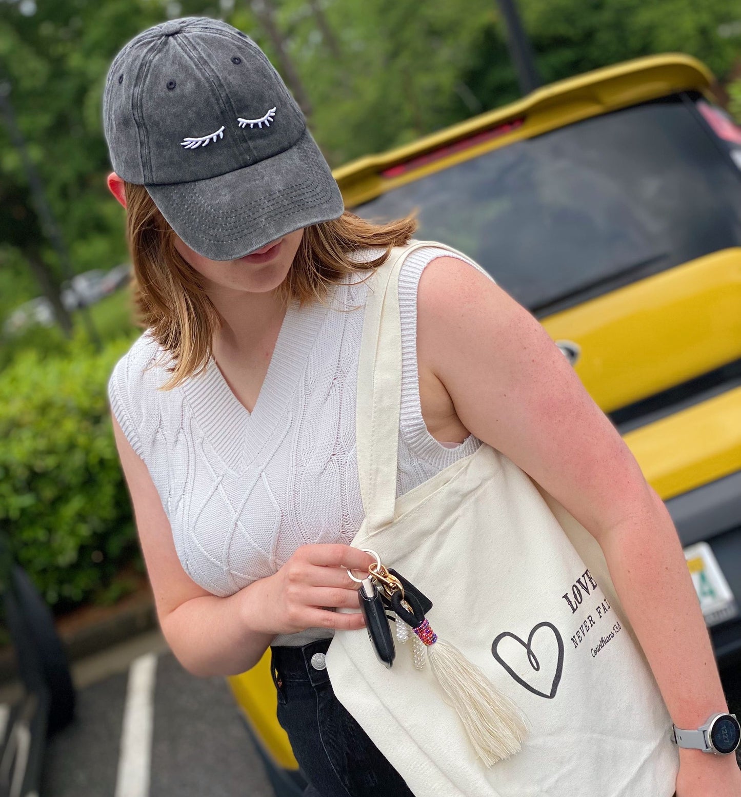 A woman wearing a gray baseball cap and a knitted white sleeveless top holds a cream tote bag with a heart graphic, showcasing colorful tassels and car keys, with a bright yellow car in the background.