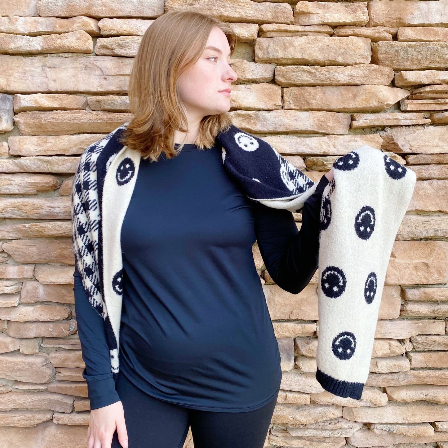 Casual outfit featuring a long-sleeve black top and a stylish patterned scarf, standing against a textured stone wall.