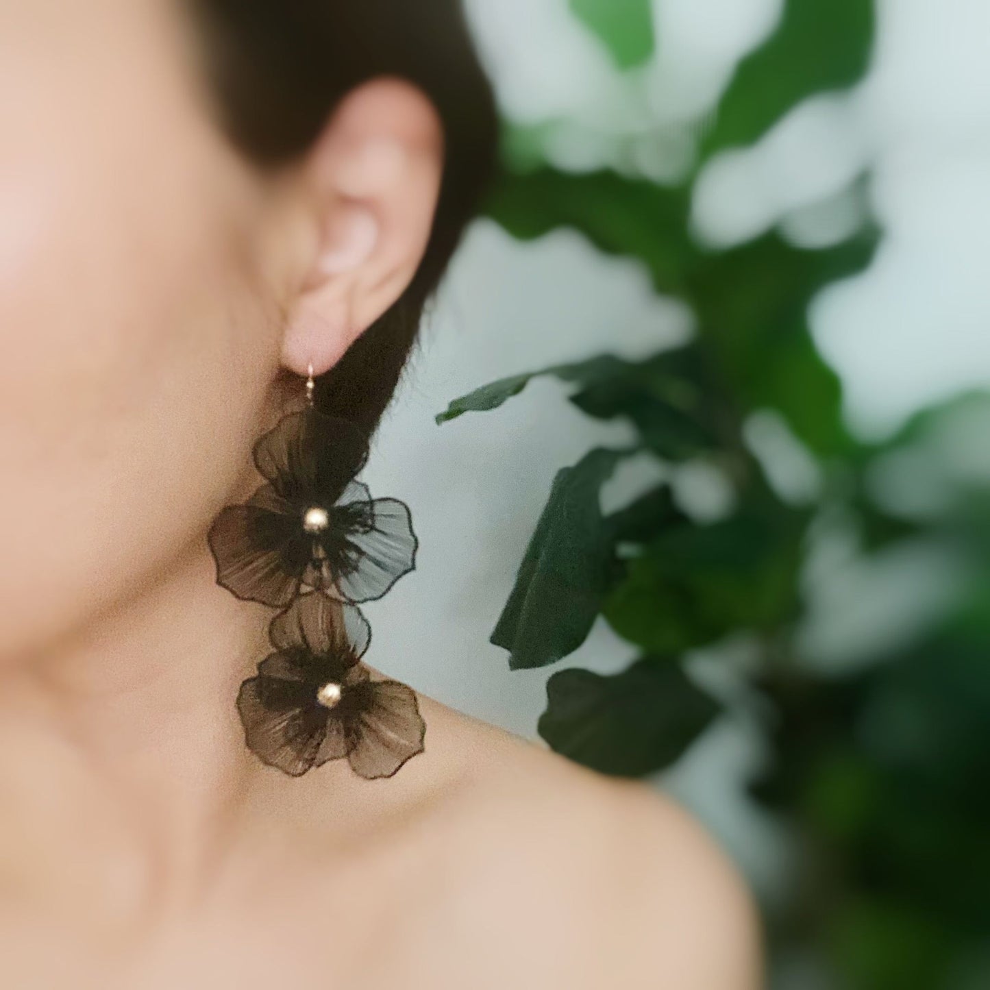 Elegant black floral earrings with pearl accents, worn by a woman with a simplistic background featuring green foliage.