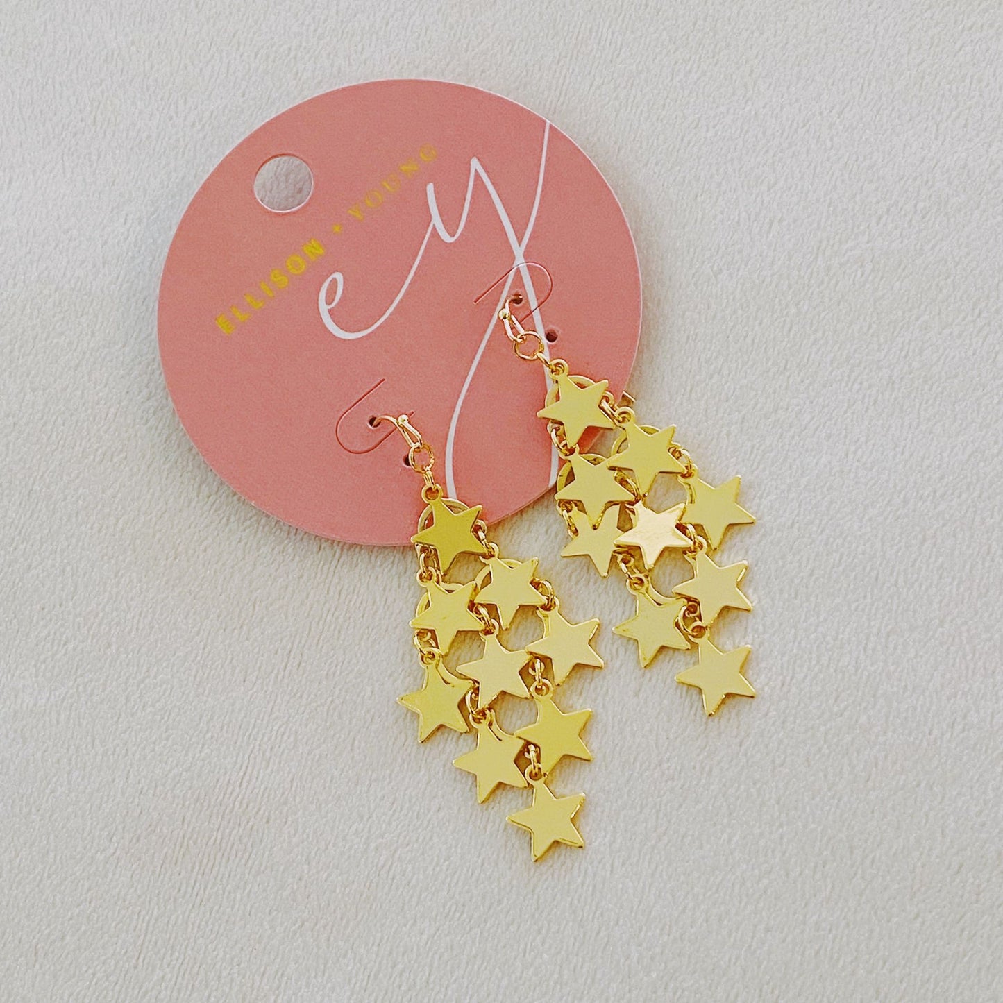Gold star dangle earrings on a round pink display card, featuring multiple shiny star charms cascading in a vibrant and playful design.