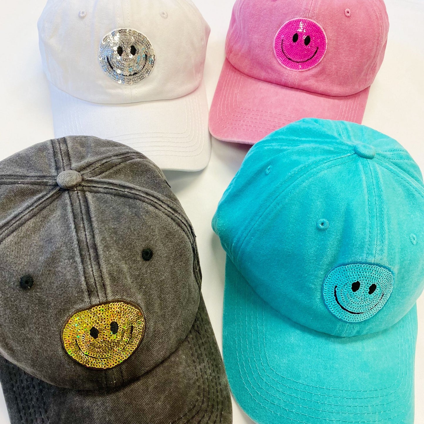Colorful assorted smiley face hats featuring sequined designs, including pink, turquoise, white, and gray caps, arranged on a white background.