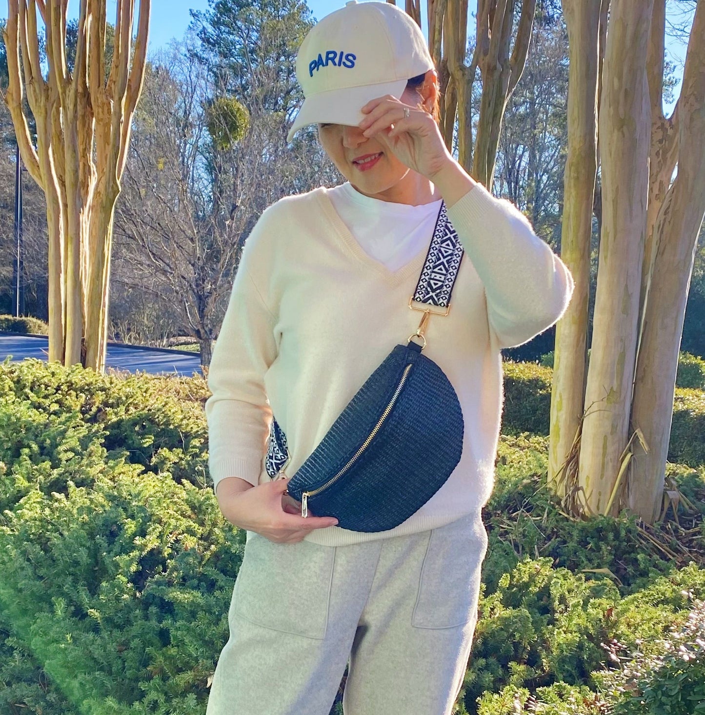 Stylish woman wearing a light sweater and gray sweatpants, holding a trendy black crossbody bag with a patterned strap, set against a scenic outdoor background with greenery and trees.