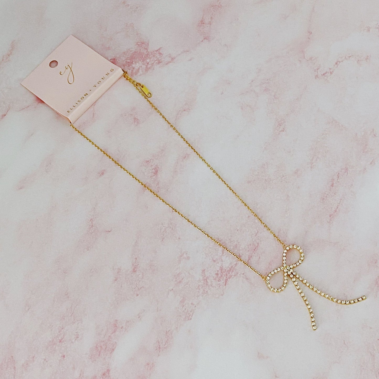 Gold necklace with a sparkling rhinestone bow pendant, elegantly displayed on a pale pink marble background.
